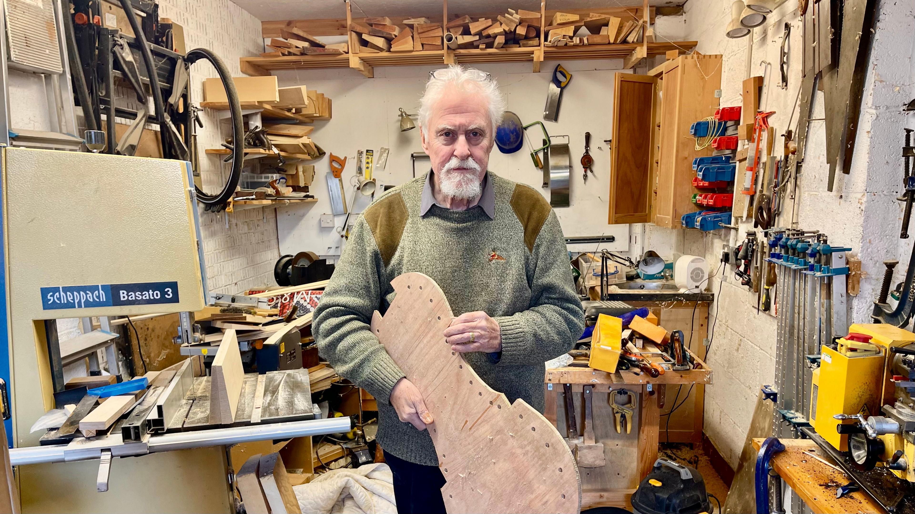 A man holds a wooden template of a cello inside a very cluttered garage. The man has short grey hair, a grey beard, glasses balanced on his head, a green jumper with brown shoulder pads and navy trousers. The garage is filled with wood, tools, machinery, a ladder and a bike wheel.