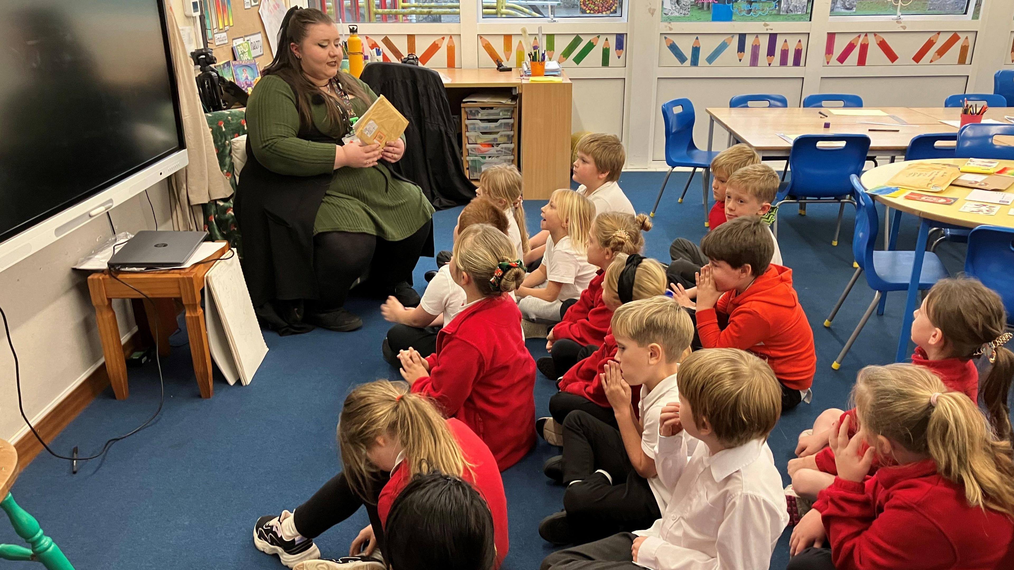 Teacher holding an envelope sitting in front of about 16 pupils who are all cross-legged on the floor in front of her in a classroom. 