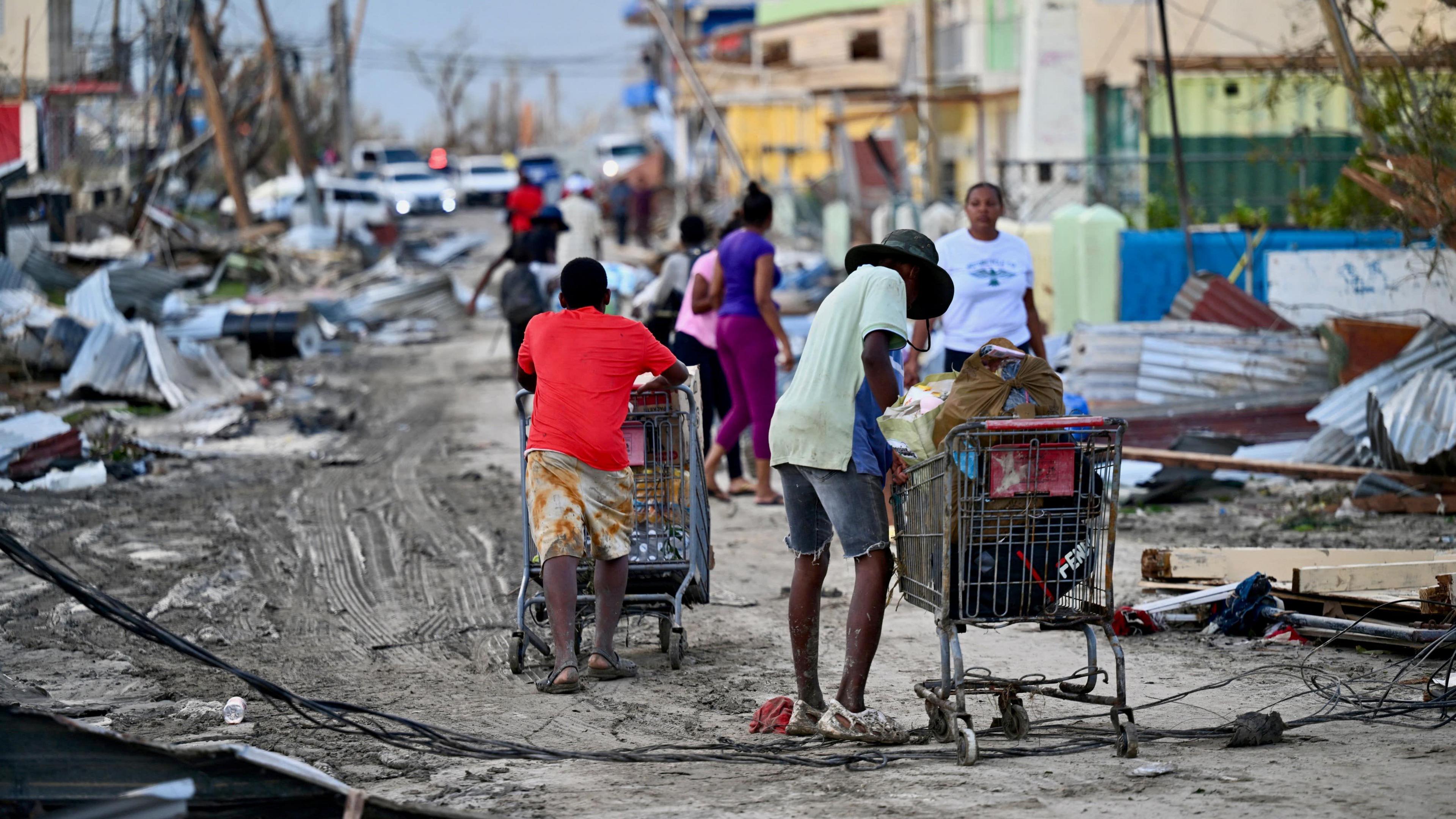 People push shopping trolleys along a road flanked by fallen debris and mess.