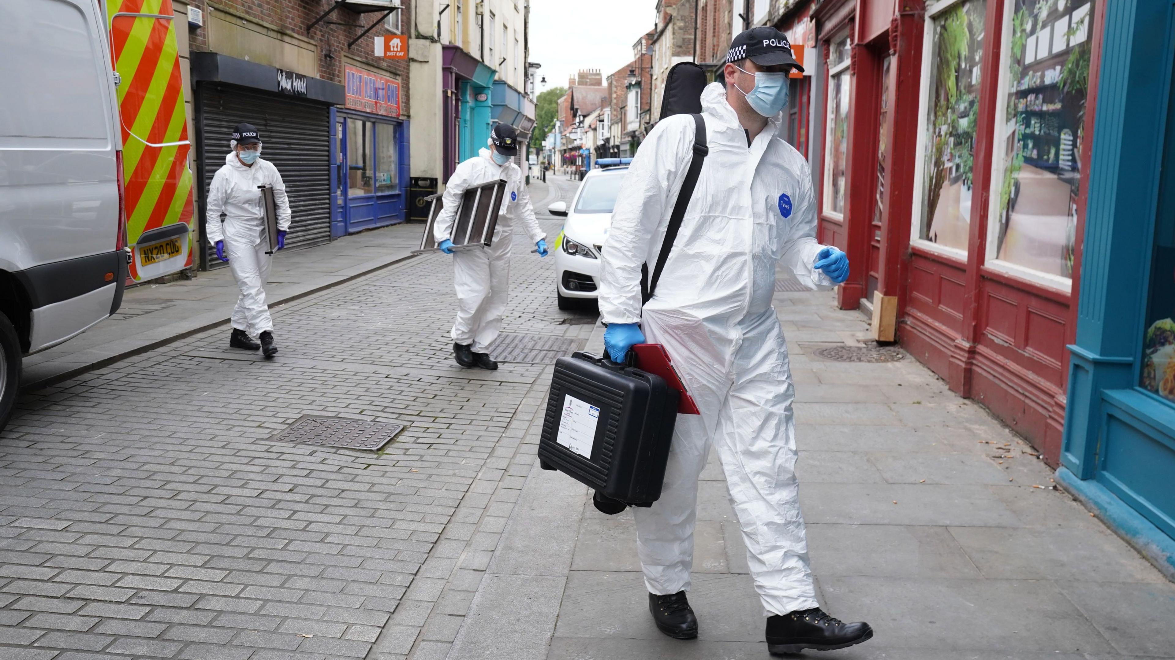 Police forensics officers wearing white bodysuits and masks carry equipment from the back of a police van towards a building just out of shot