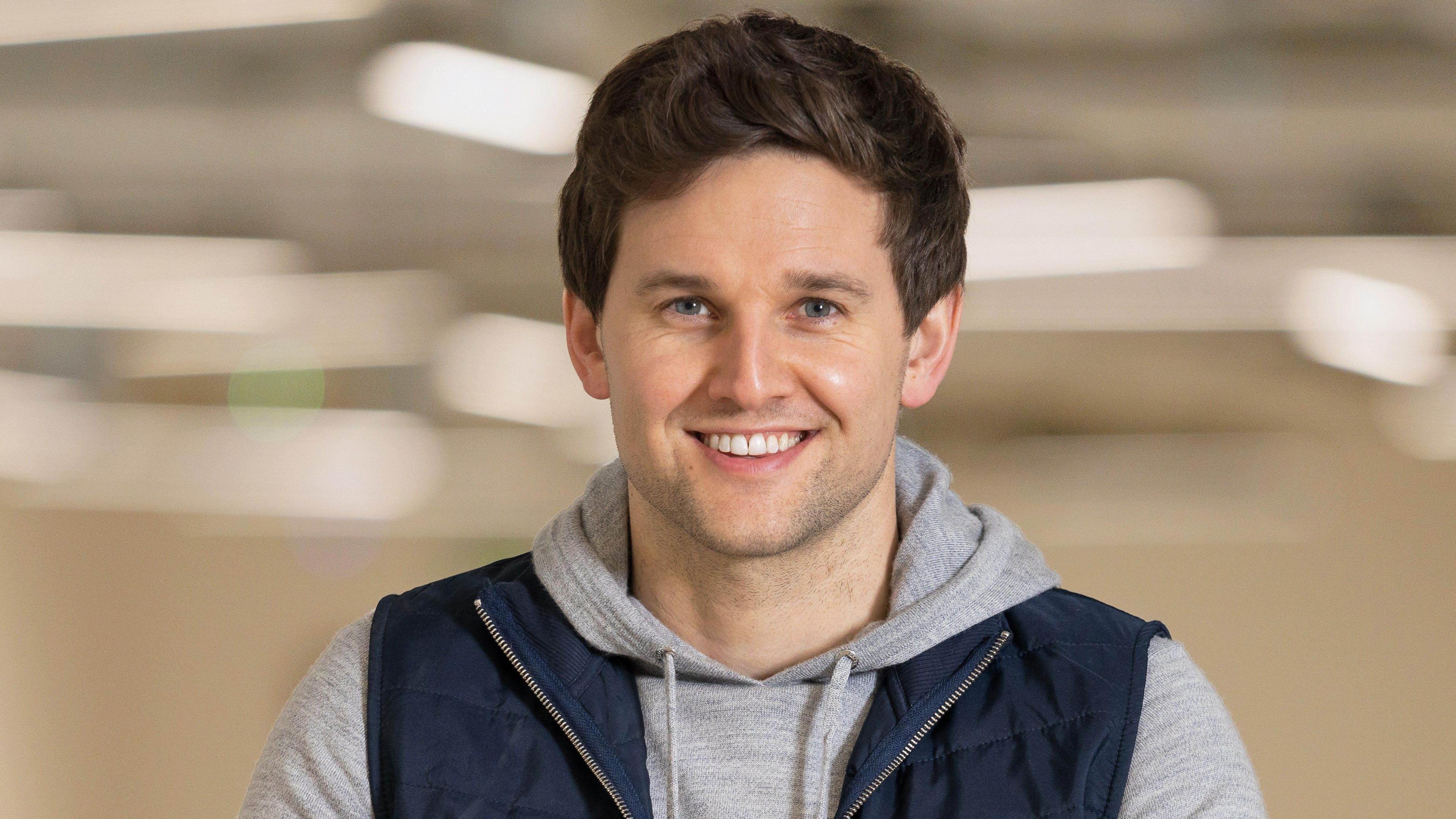 A man in his 30s smiling at the camera, he has brown hair and wears a gray hoodie and navy zip-up vest. The background is beige and blurred. It is a head and shoulders shot.