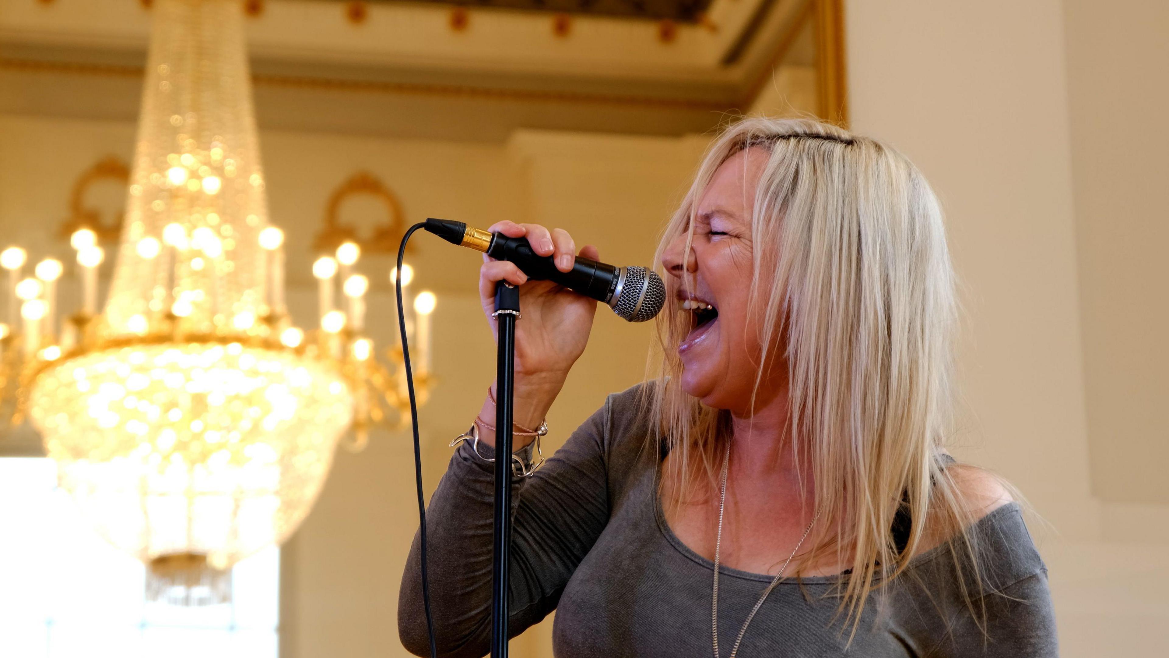 A woman with long blonde hair wearing a grey long sleeved top singing passionately into a microphone with a chandelier in the background