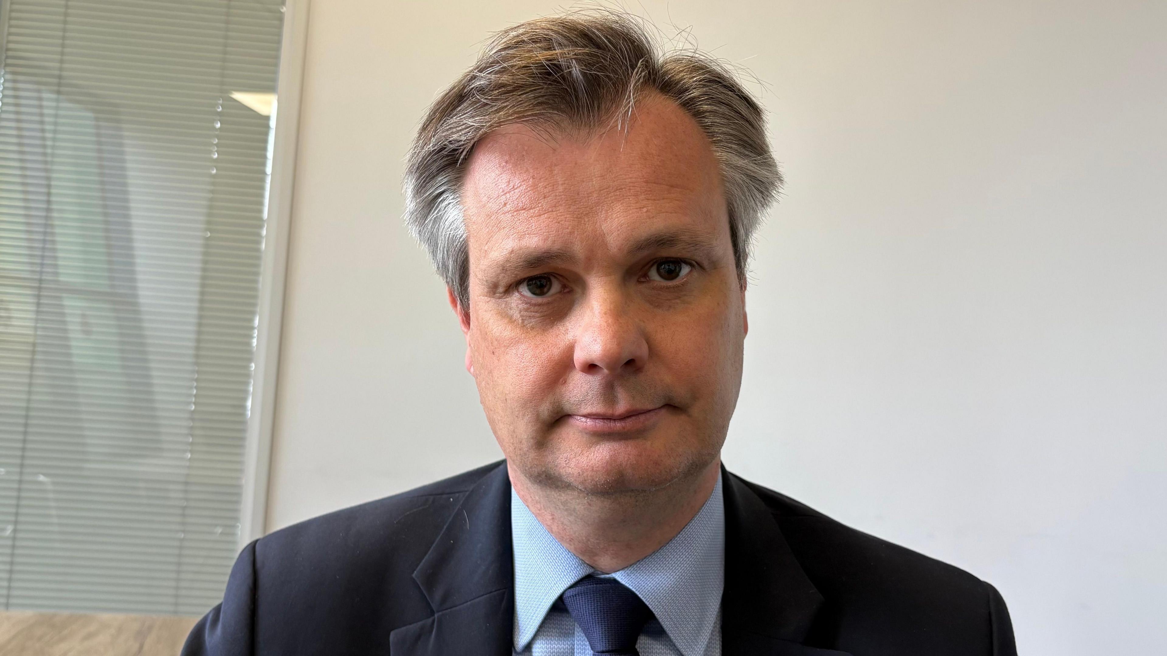Simon Rouse with medium-length dark hair combed back. He is looking at the camera and wearing a dark blue jacket with light blue shirt and dark blue tie. He is standing in front of a white wall, with a window covered by a blind to his right.