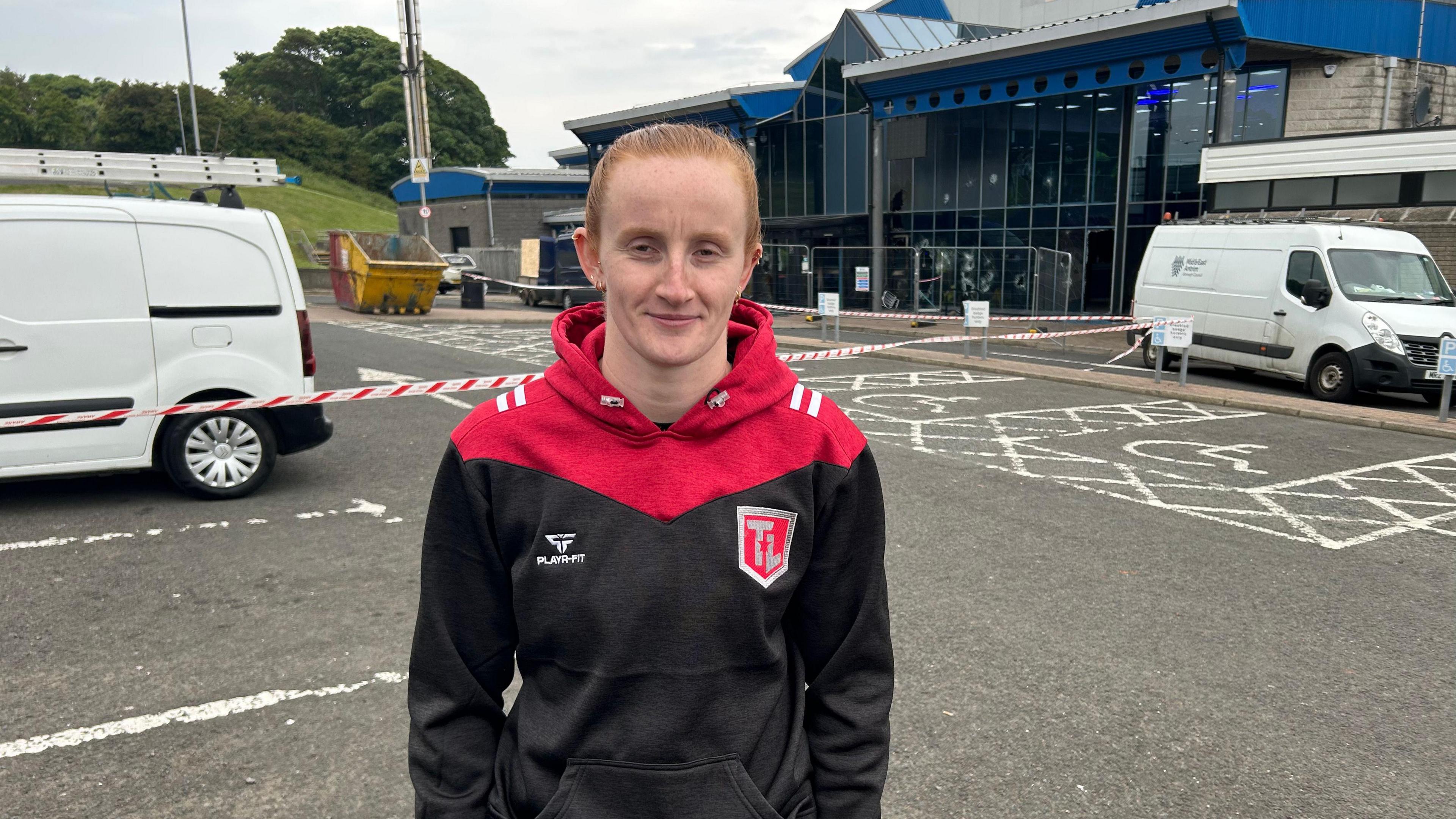 A red haired woman wears a red and black hoodie. Behind her are tow white vans either side of a glass front building with a blue roof. There is red and white tape between her and the building.