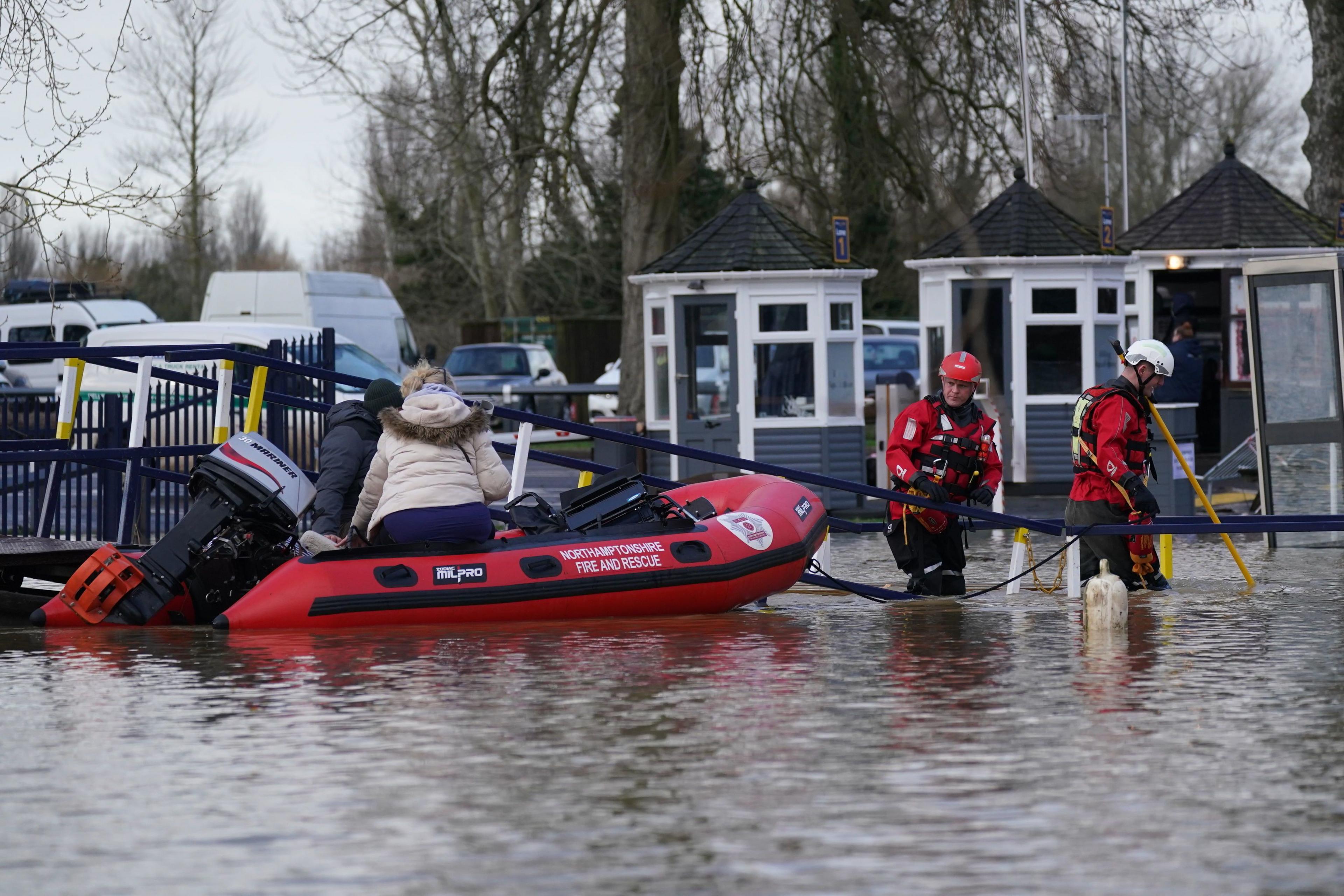 Two men from a fire and rescue service wearing red gear and helmets are wading in the flooded water and dragging a boat with two people on it. The water comes up to just above the knee level of the two men. We can not see the faces of the two people on the boat but one has a white winter coat and has blond hair tied at the back.