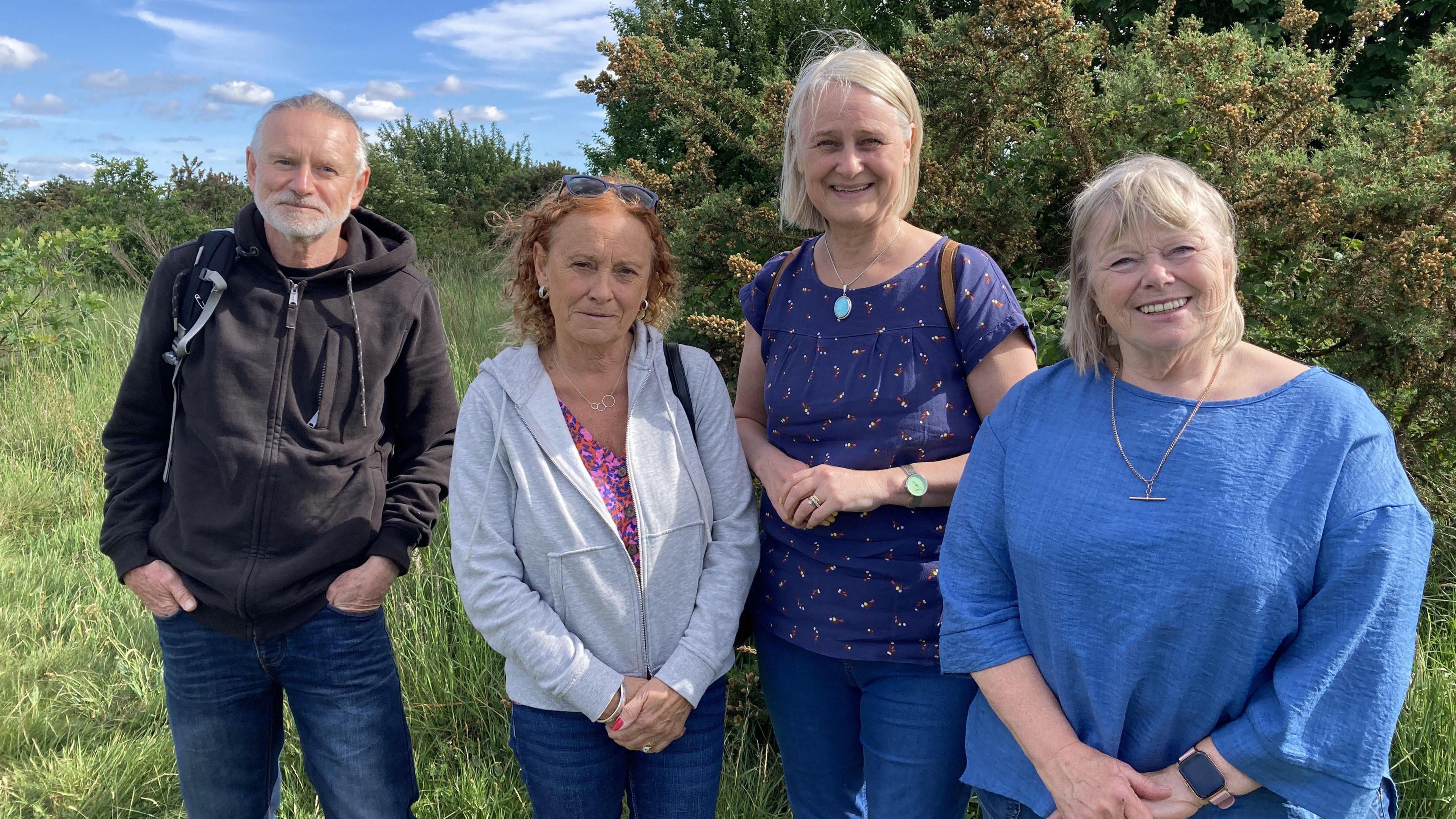 Four people stood on a large grass field, smiling - including a man wearing a black jumper, a woman wearing a white jumper and sunglasses, another woman wearing a blue T-shirt, and another woman wearing a blue top. Behind them are a few trees.