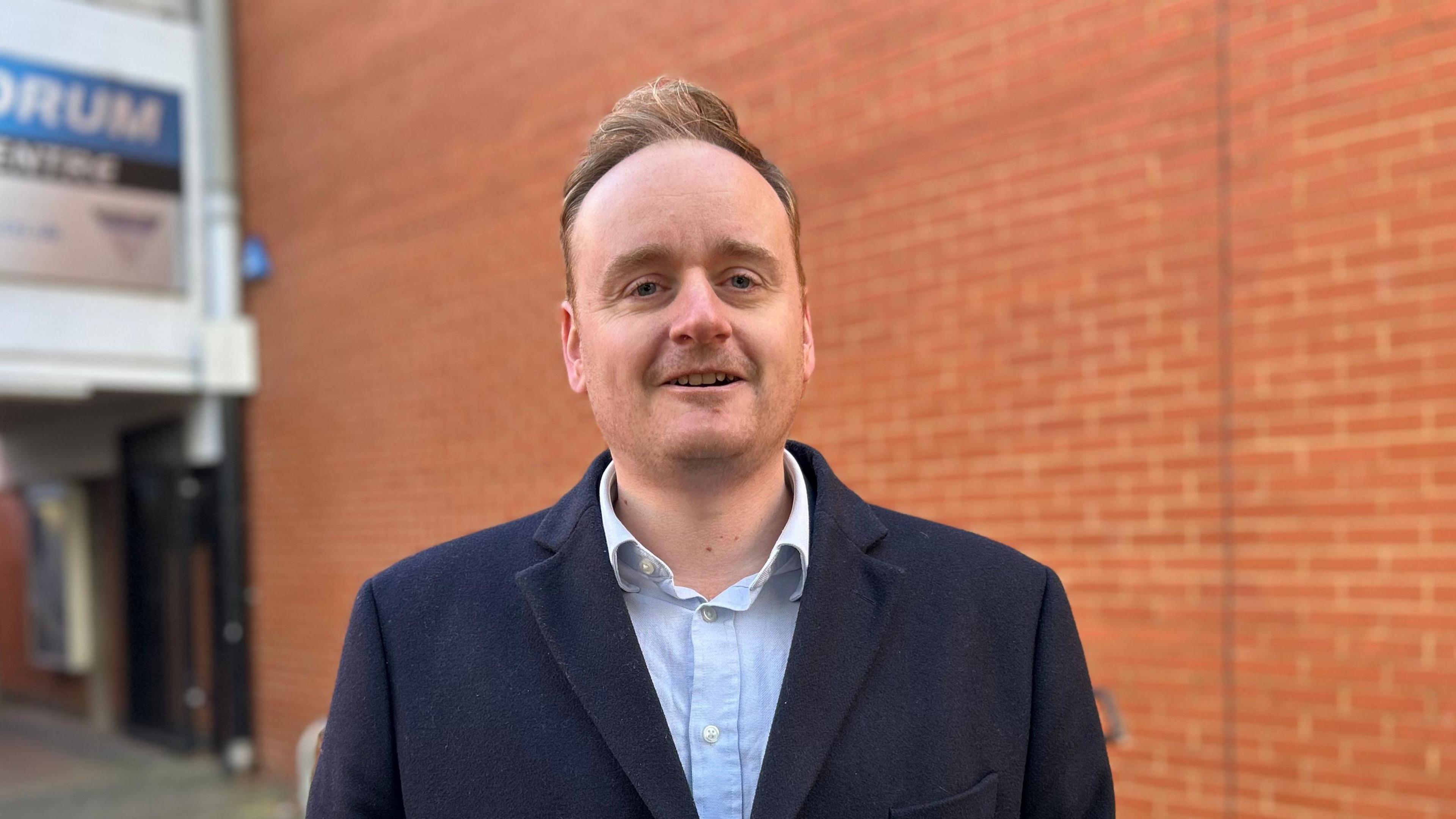 Adam Brown wearing a blue blazer and white shirt. He is stood in front of a red brick wall and looking directly at the camera. 