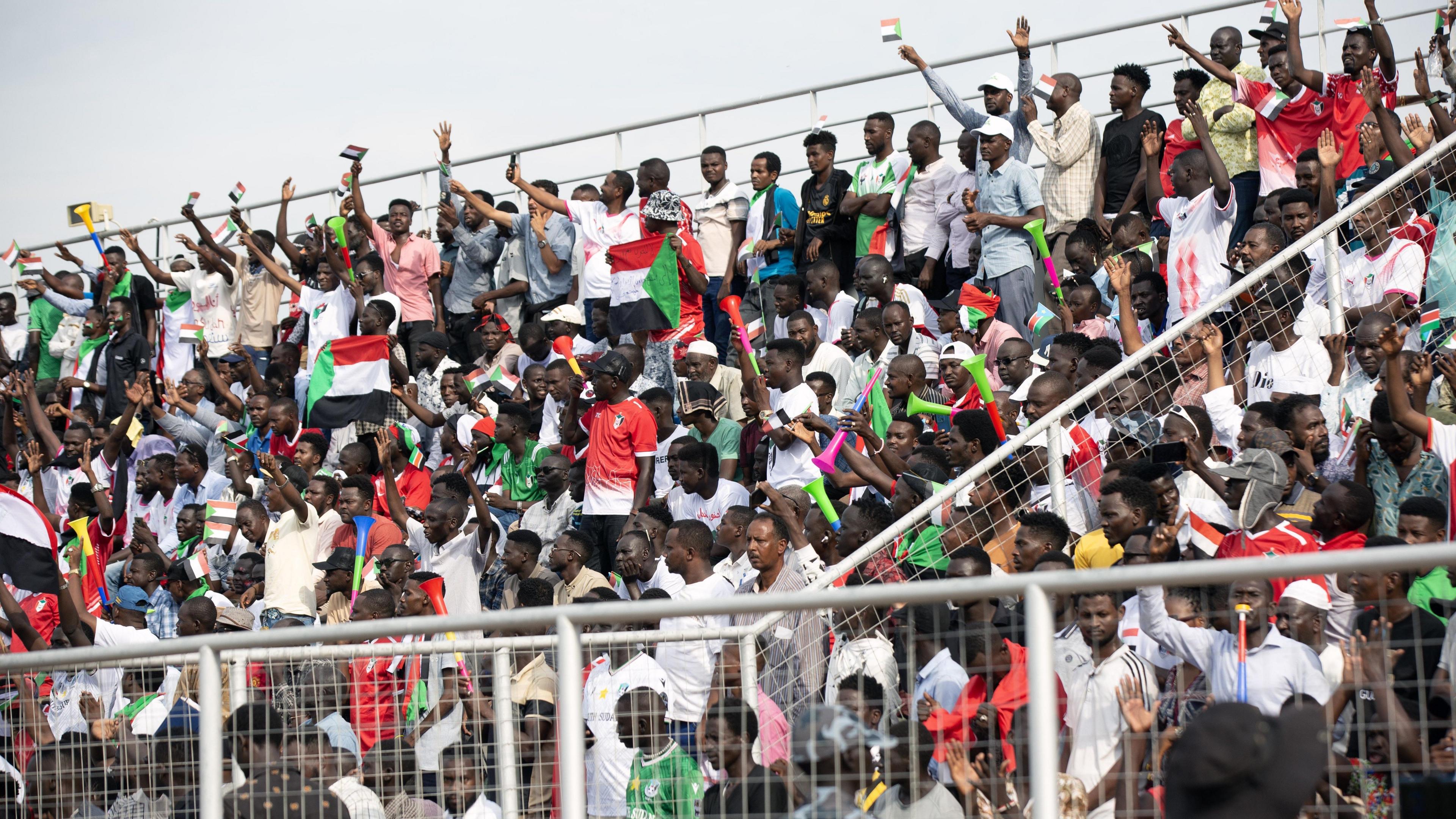 A packed stand shows Sudan fans waving flags, with their arms in the air and cheering in Juba National Stadium