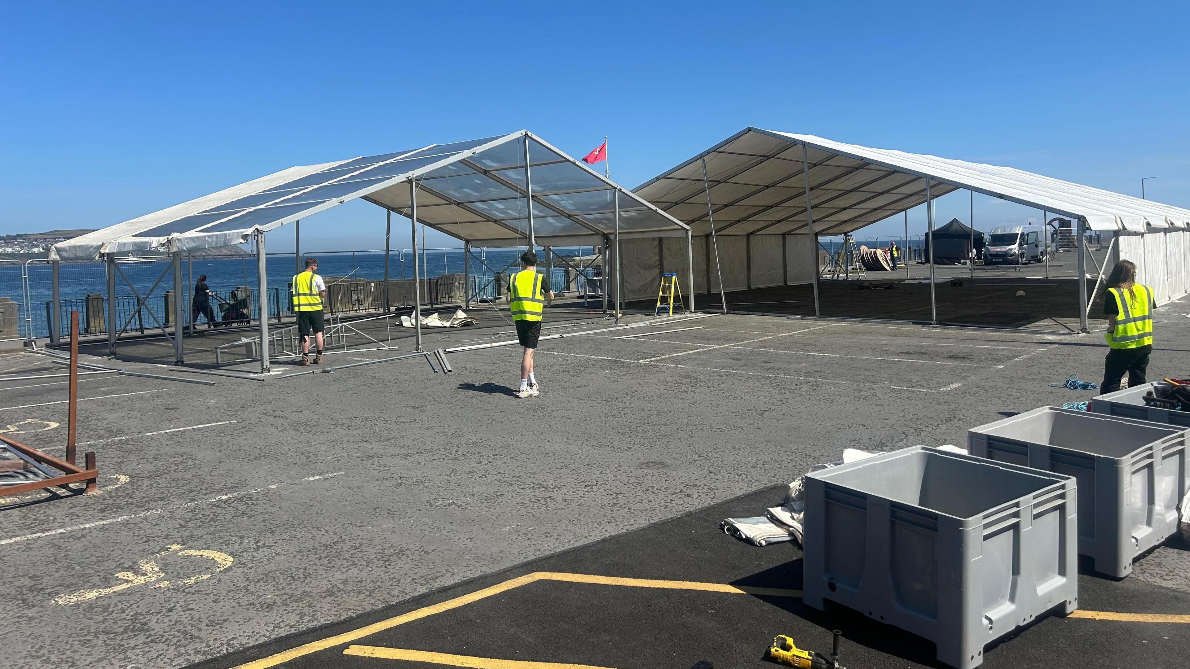 The car park space has two large marquee structures being built, one has a white covering over its roof and two sides. Three people in hi-vis jackets work on the site. You can see the sea in the distance.