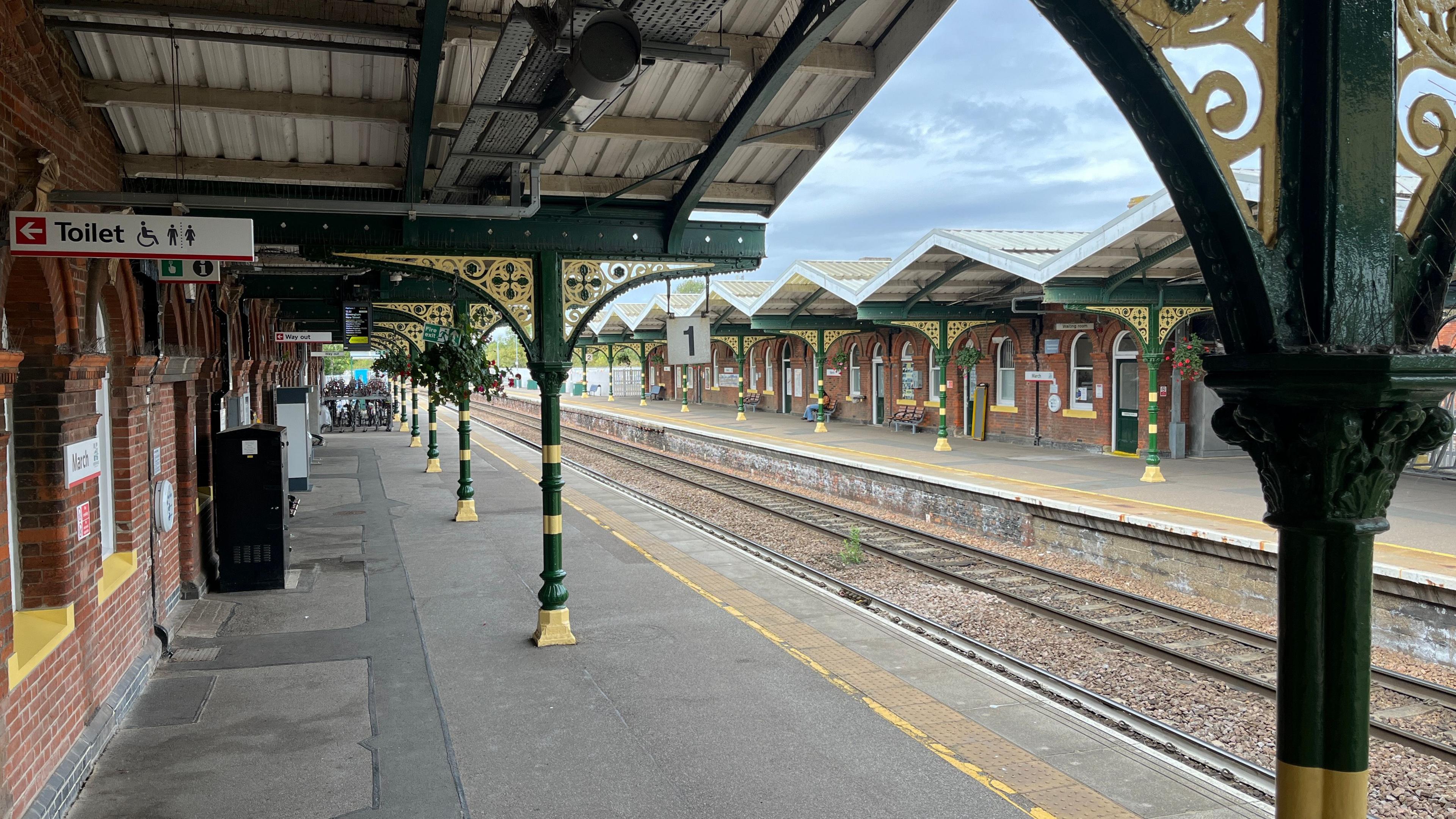 Two platforms on a Victorian railway station have ornate yellow and green pillars and arches made of iron. There are modern-day signs for passengers and train tracks between the two platforms
