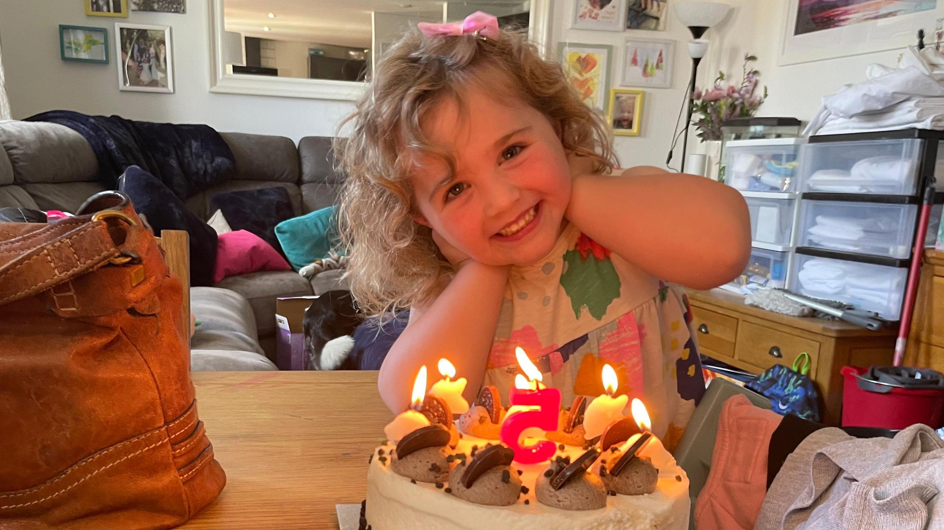 A little girl is smiling, she has a birthday cake in front of her with candles and the number 5. 