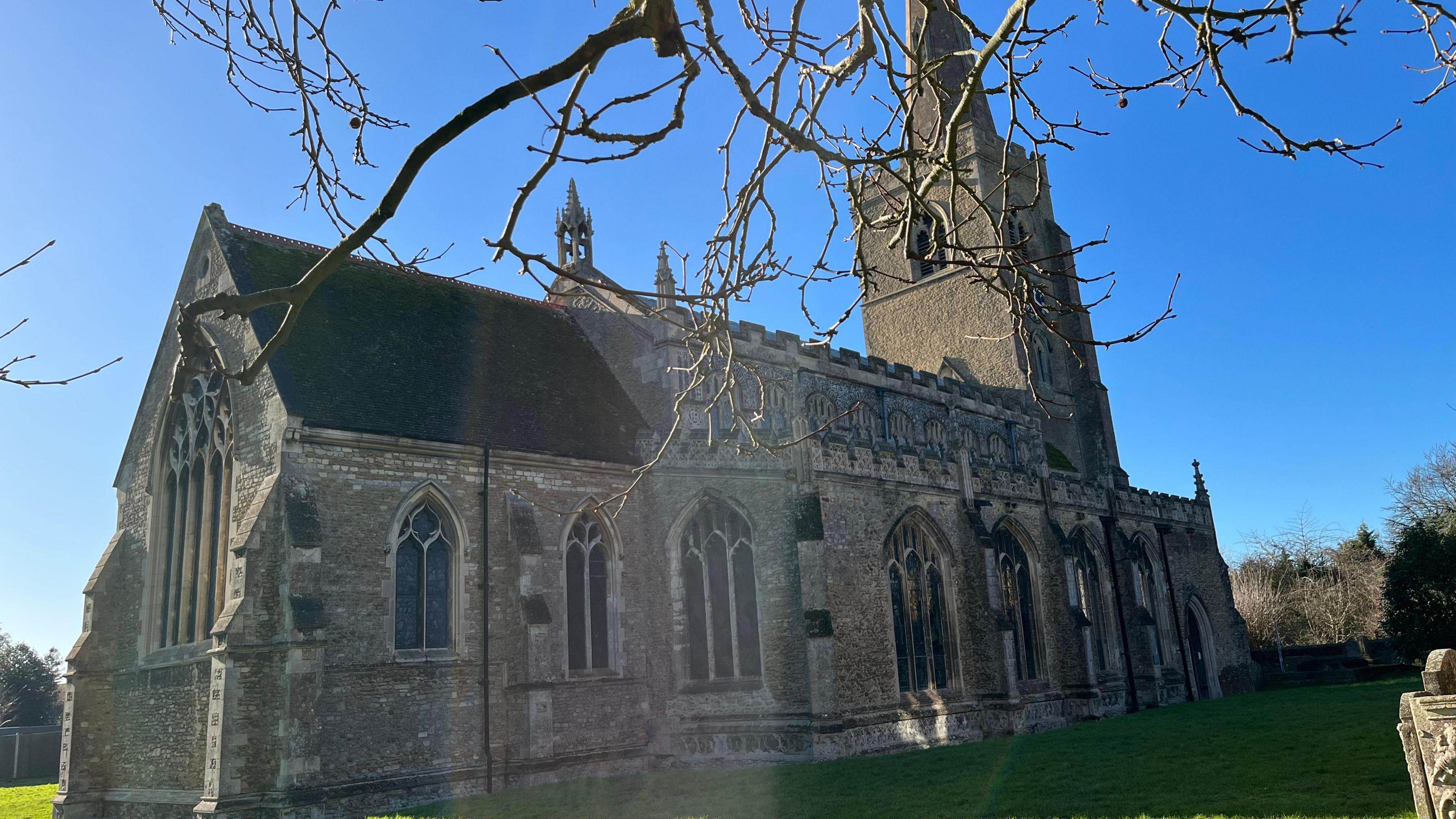 The outside of St Wendreda's Church in March. There is a blue sky and the spire can be seen at the far end of the building, with tree branches in the foreground.