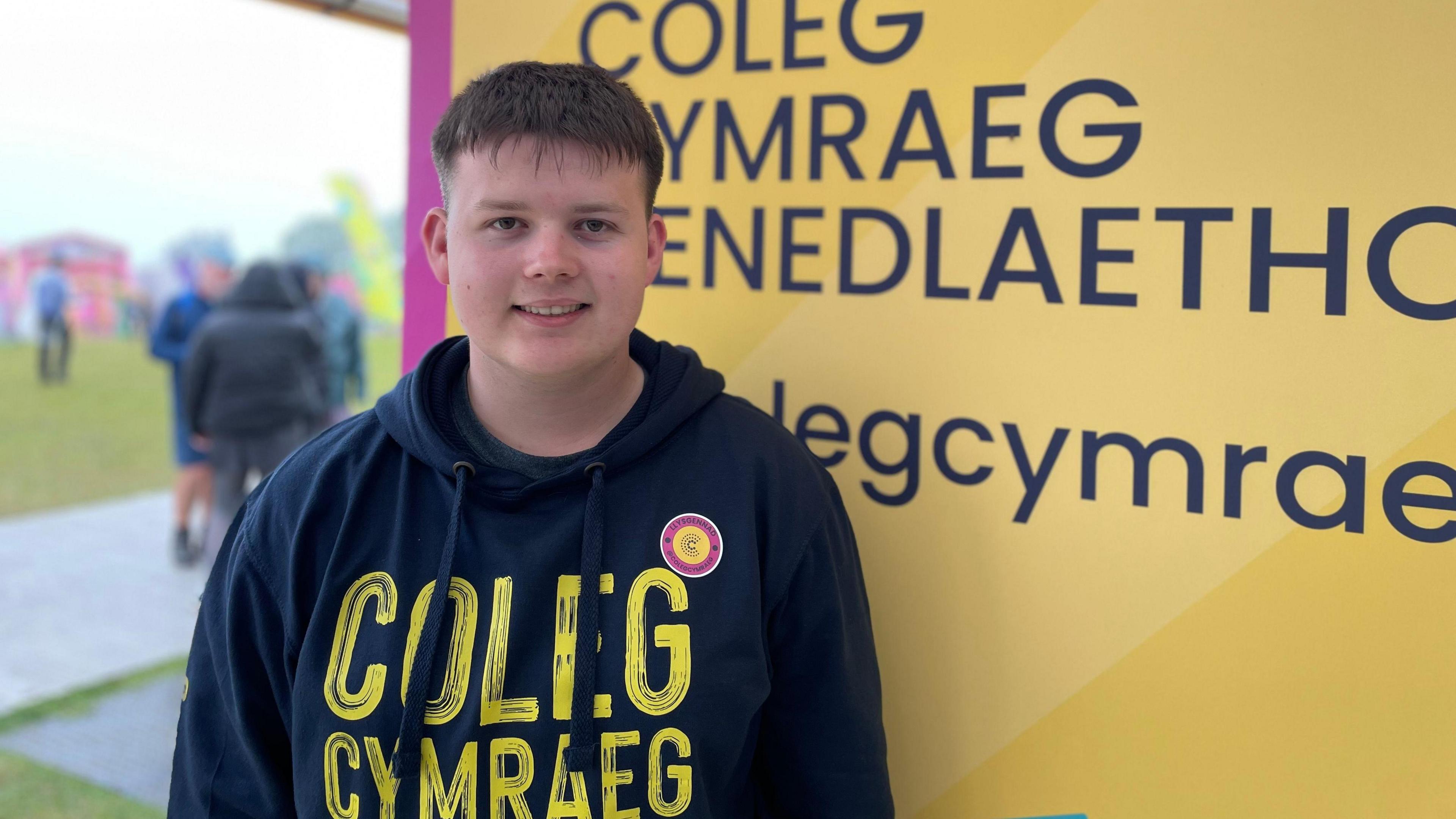 19-year-old Iwan wears a navy pullover hoodie which reads "Coleg Cymraeg" in yellow font. He smiles at the camera showing his teeth and has light brown short hair.