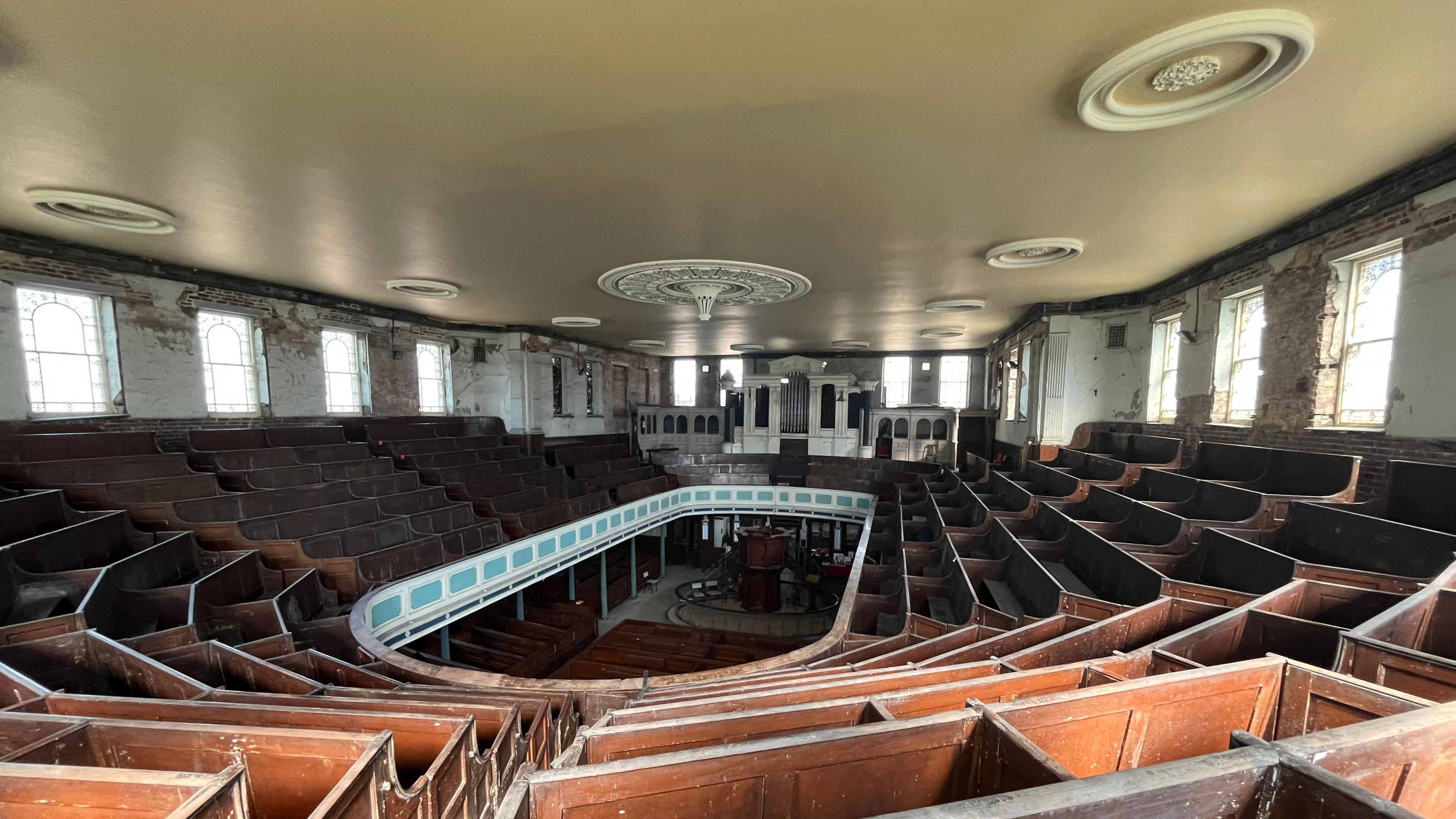 The inside of a Methodist chapel, viewed from the upper floor which is filled with brown pews where worshippers sit. An oval-shaped opening is visible in the centre of the room where the floor below can be seen. A church organ is visible at the far end of the room.