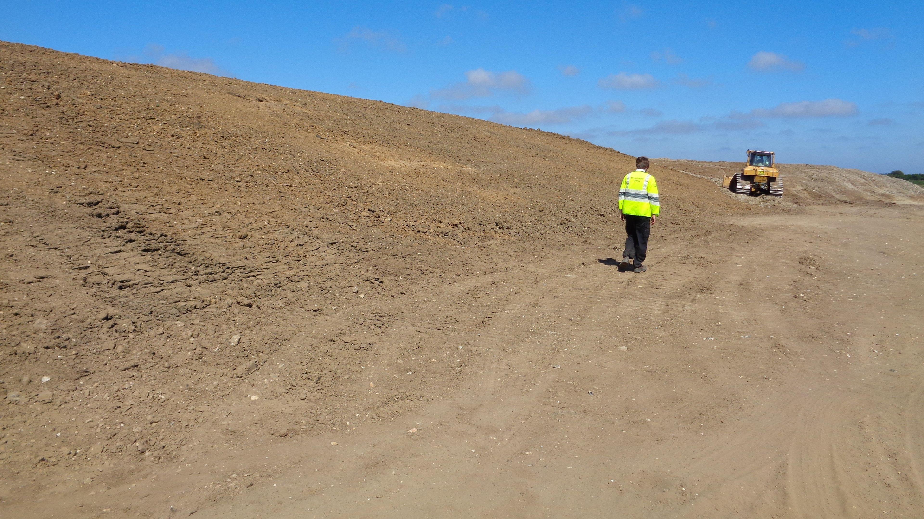 Large mounds of dirt  on the left of the image. In the middle of the image is a person with a fluorescent jacket, in the background is a yellow machine digger.