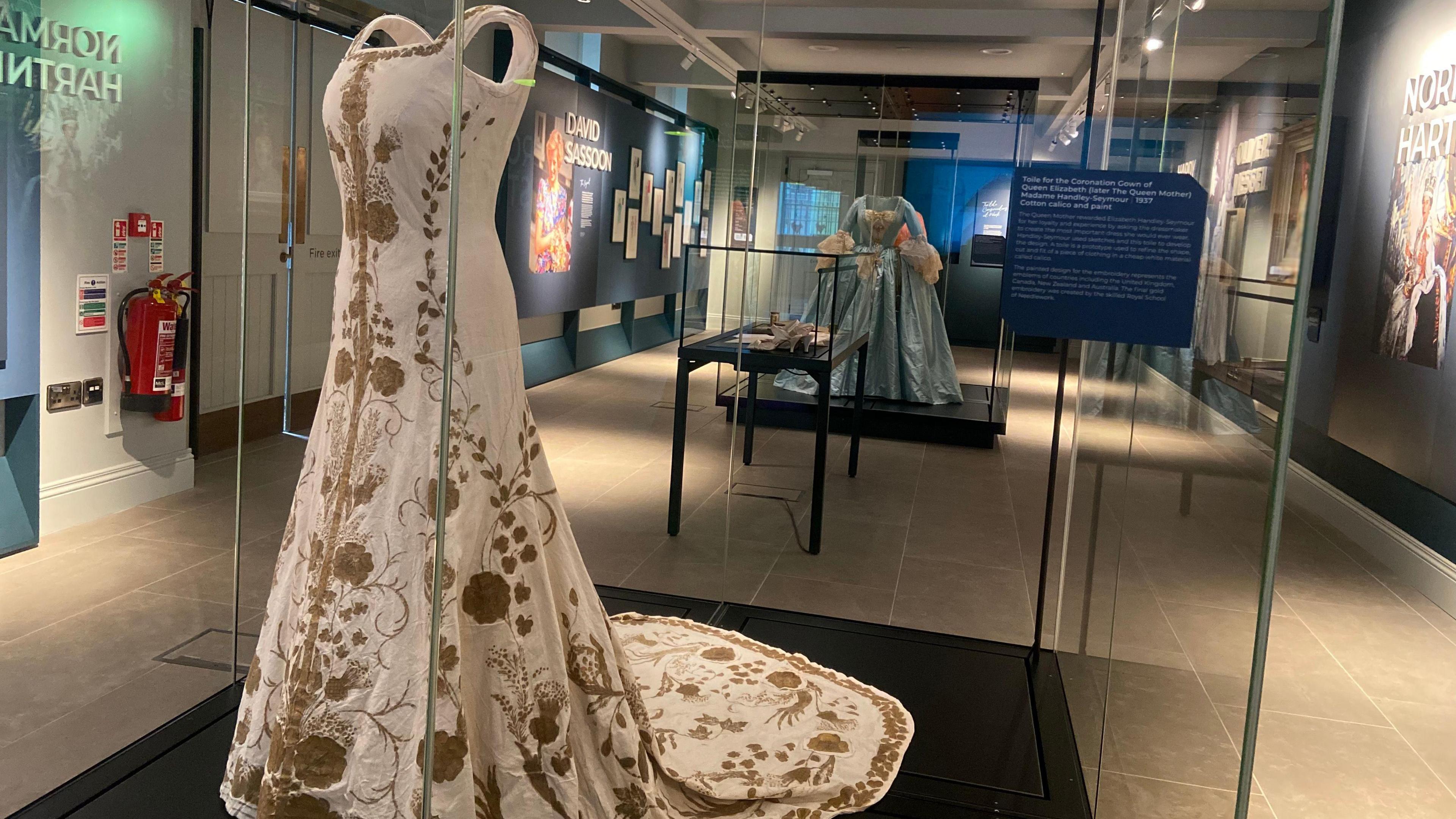 Toile for the 1937 coronation gown of Queen Elizabeth, later the Queen Mother. It is a full length white gown with gold embroidery and a small train. It is encased in a glass cabinet.