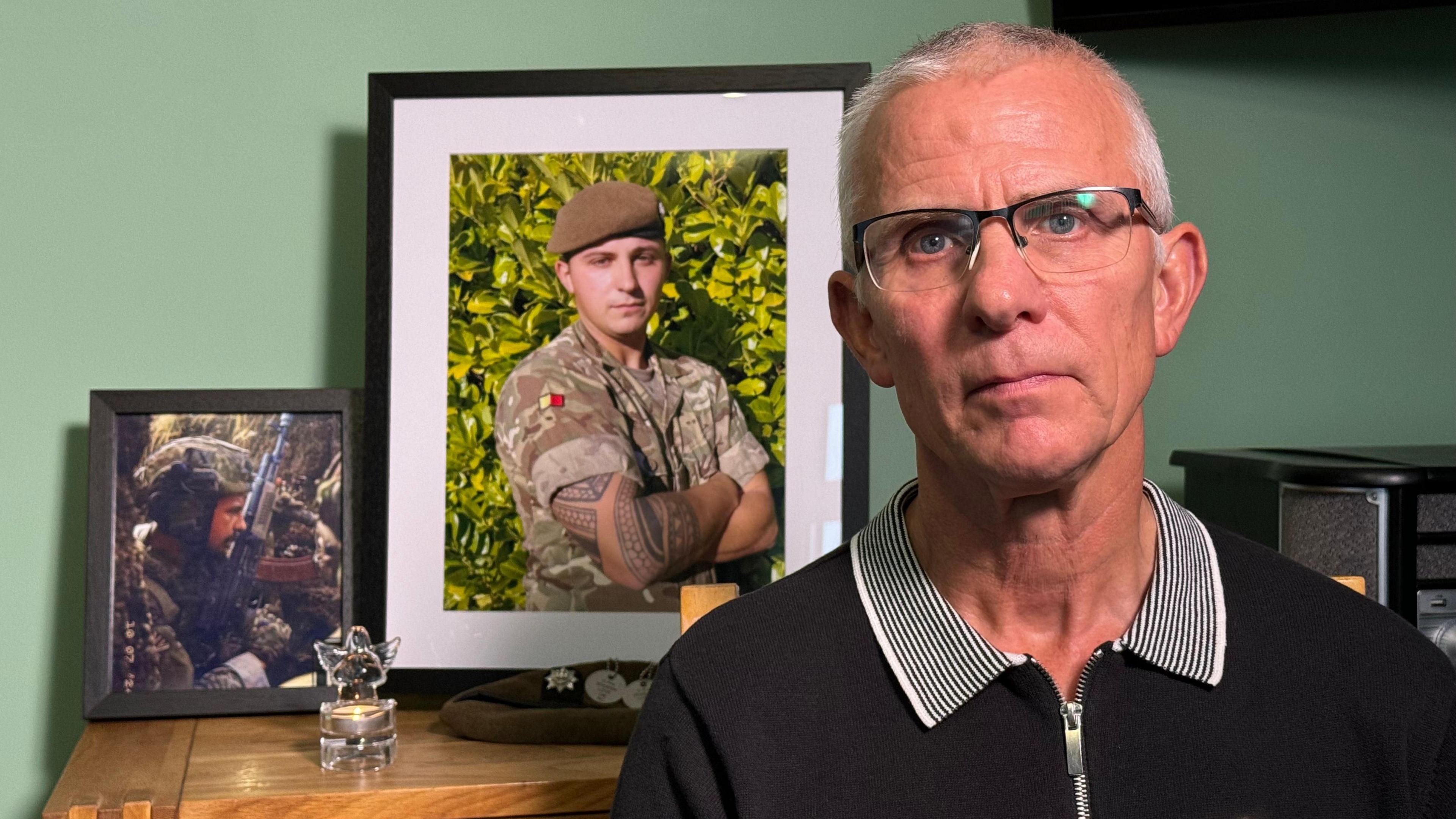 A man looks into the camera. Photos of a soldier and an ornament of an angel are sitting on a table behind him.