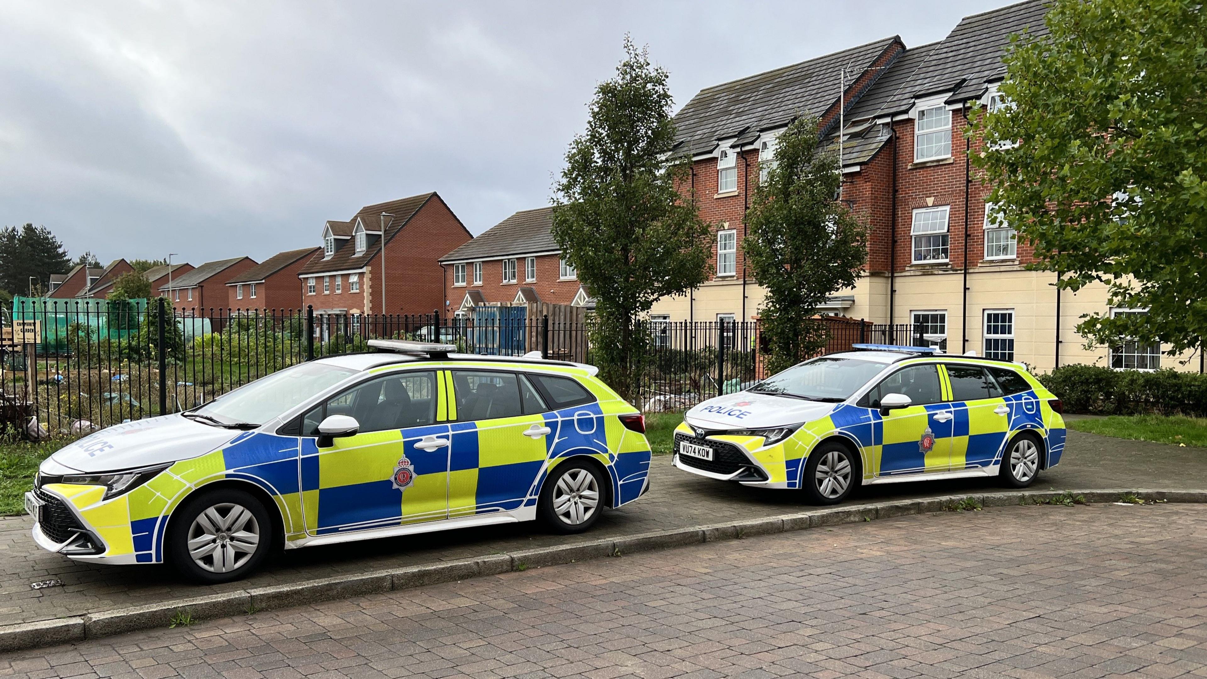 Two police cars with hi-vis yellow and blue decals, parked on the pavement on a residential estate. There is an allotment patch behind a black metal fence in the centre of the estate. In the background there is a row of flat buildings with a red brick façade on top. It is an overcast cloudy day. 