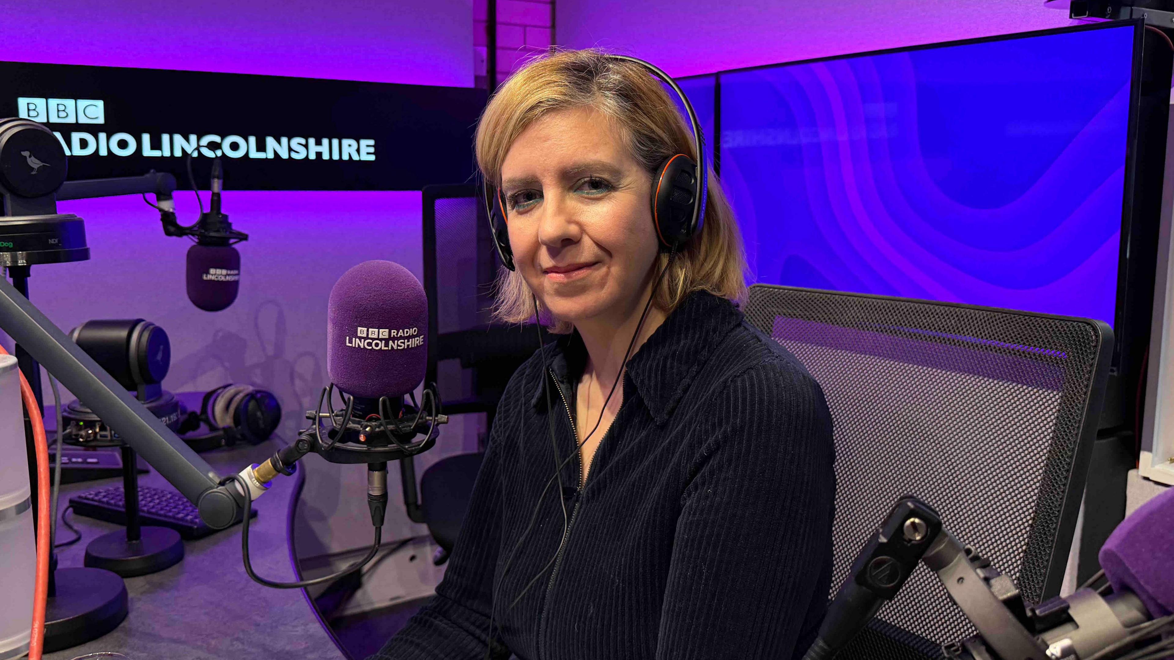 A woman with shoulder length blond hair and wearing a black top smiles into the camera. She is in a radio studio surrounded by purple lighting and video screens. She is sitting in front of a microphone with the words "BBC Radio Lincolnshire" on it.