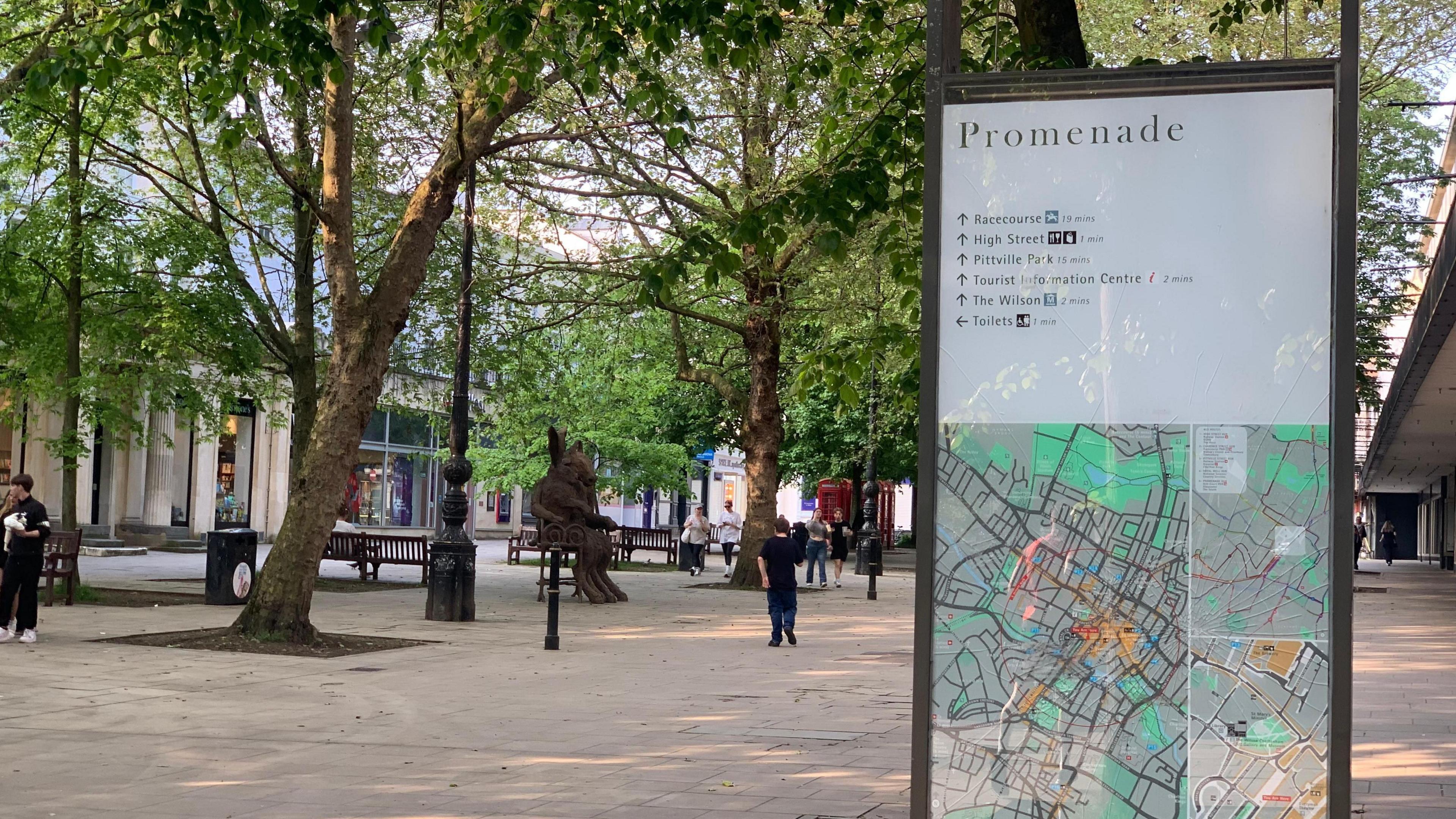 A view across the Cheltenham promenade with a large map sign in the foreground. In the background you can see shoppers walking along the flagstones, and the street is lined with mature trees. benches and statues. 