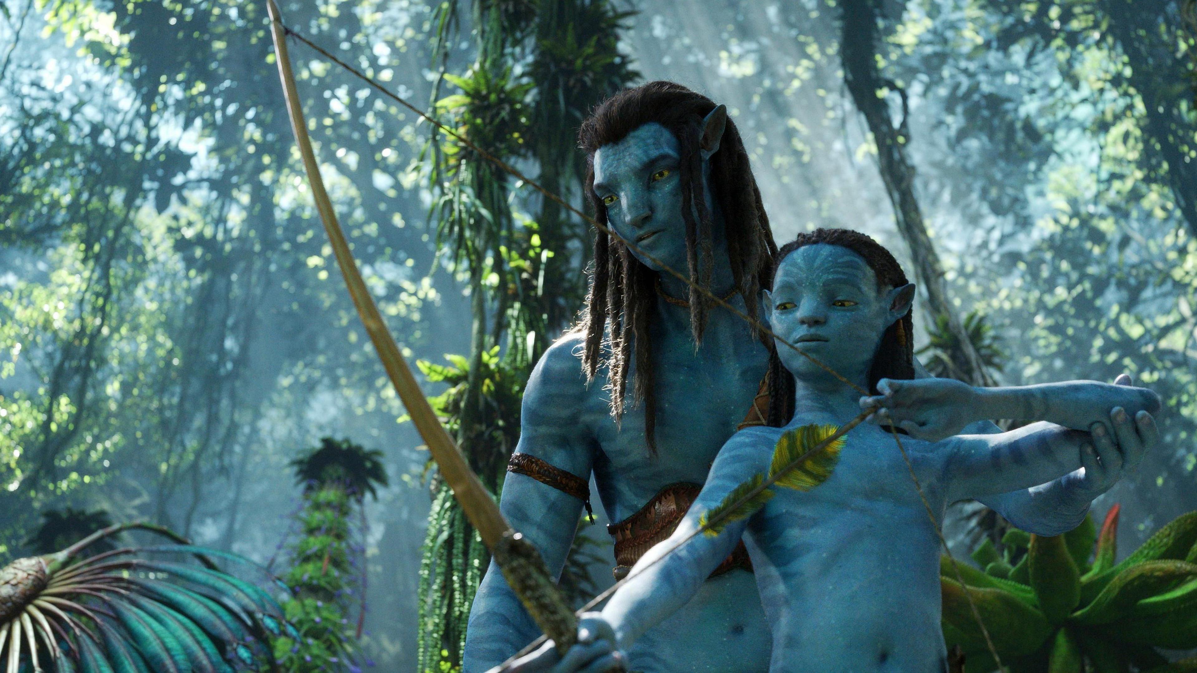 A still from the 20th Century Studios film Avatar: Way of the Water showing an adult avatar training a child avatar how to use a bow and arrow. The avatars are in the jungle. 