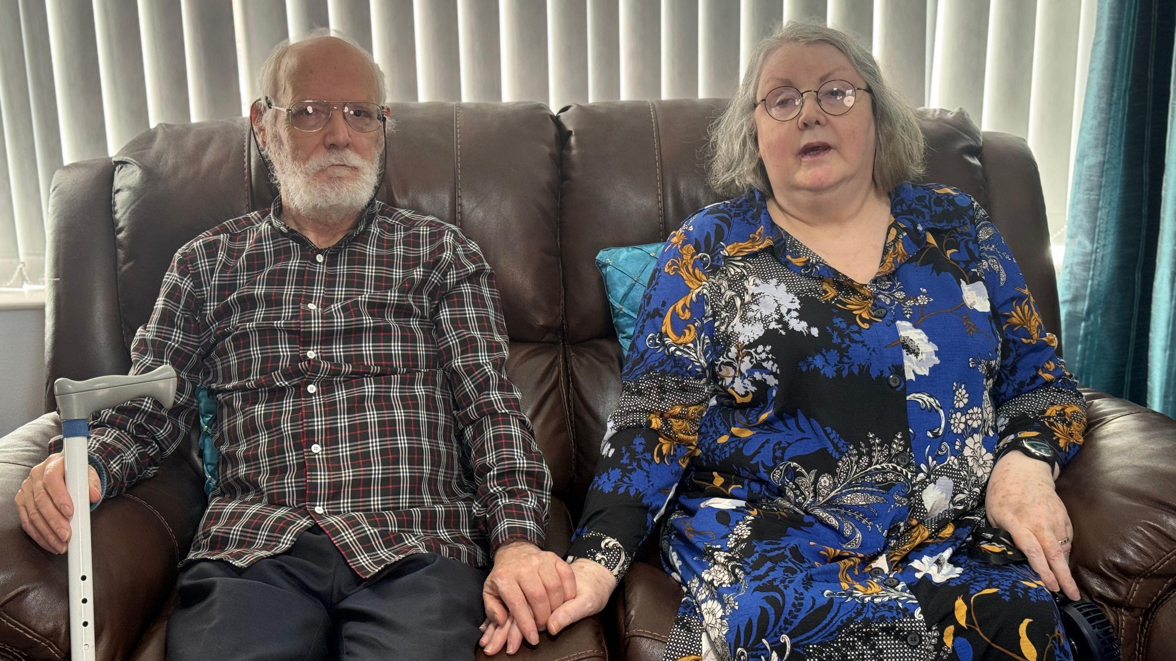 Paddy and Breda Murphy sitting on a brown sofa, holding hands. Paddy is holding a walking stick