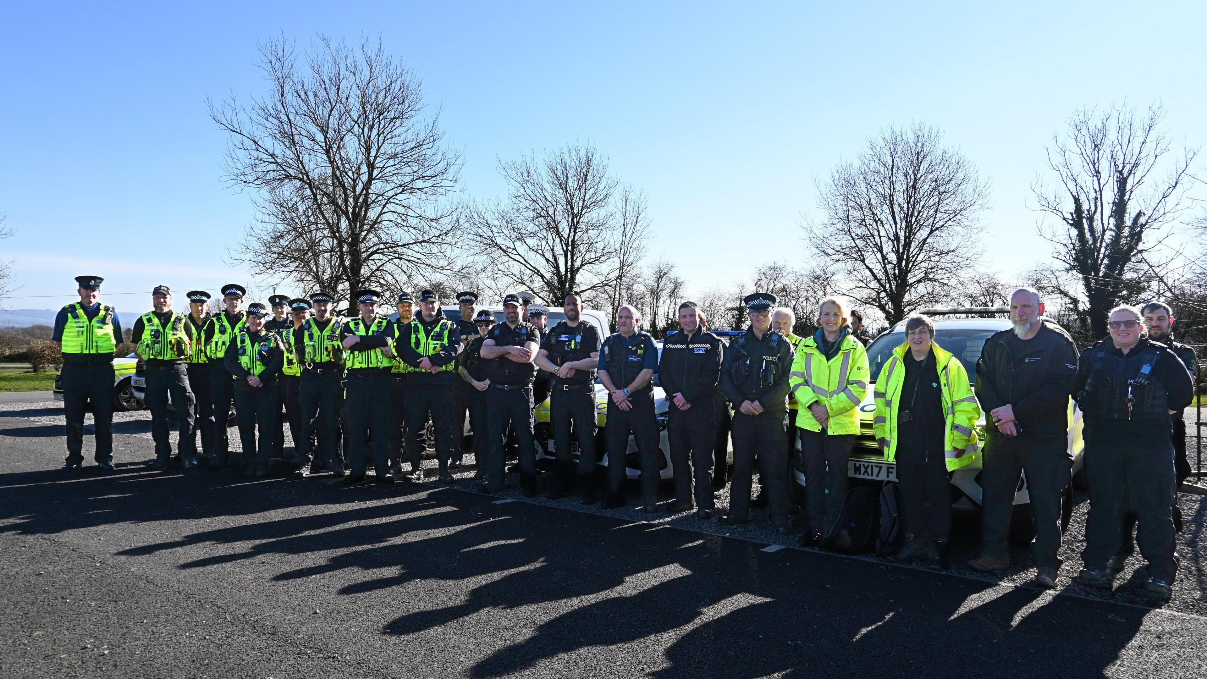 A large group of police officers standing outside in a row in front of some police vehicles