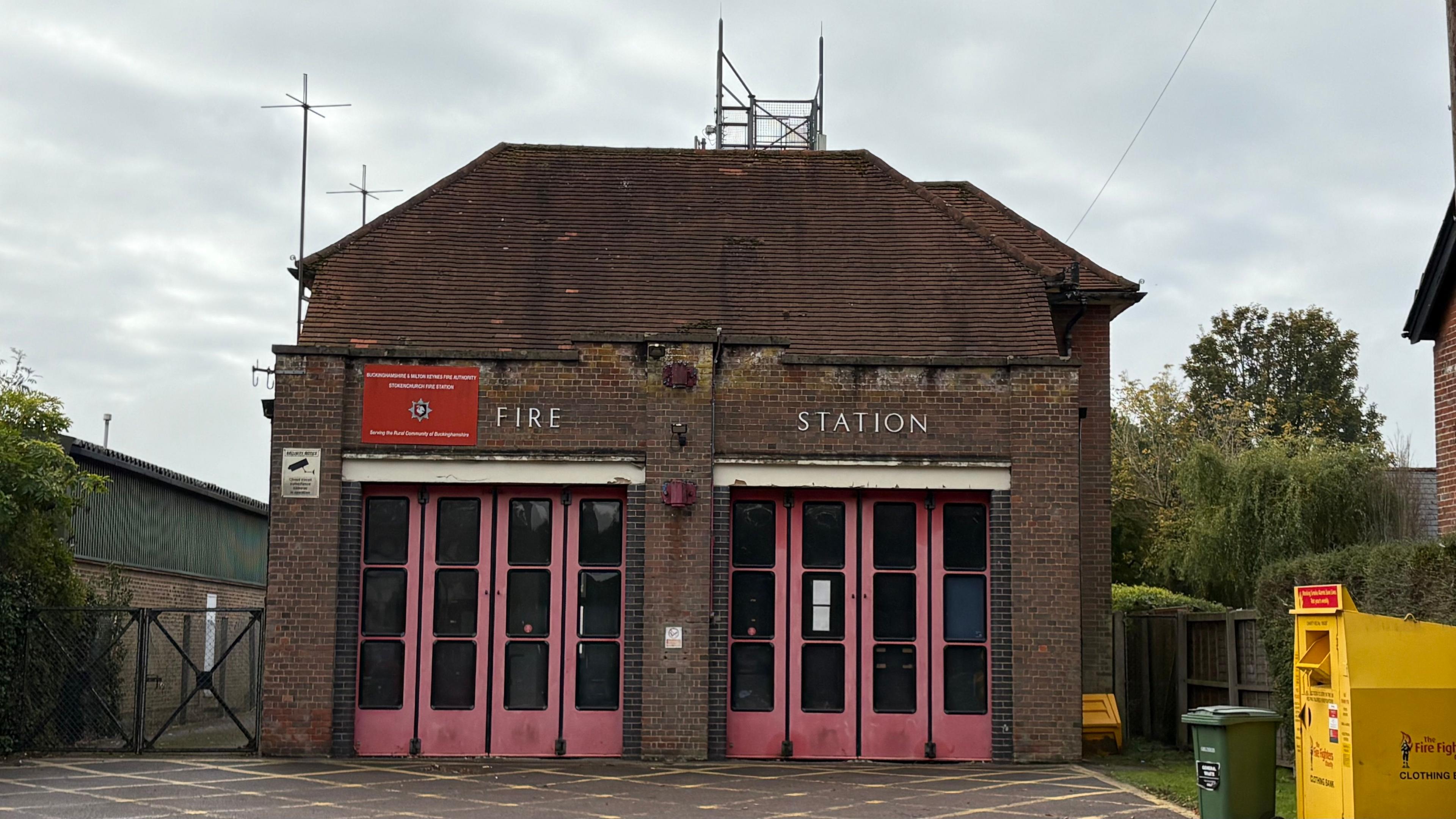 Two-storey brick-built fire station with large folding doors across the front (to accommodate fire engines). The words FIRE STATION are visible in white lettering above the doors. There are aerials on top of the building. A gate to the right gives access to the yard behind the building. The area in front is painted with yellow hatching and there is a yellow clothes donations skip to the right.