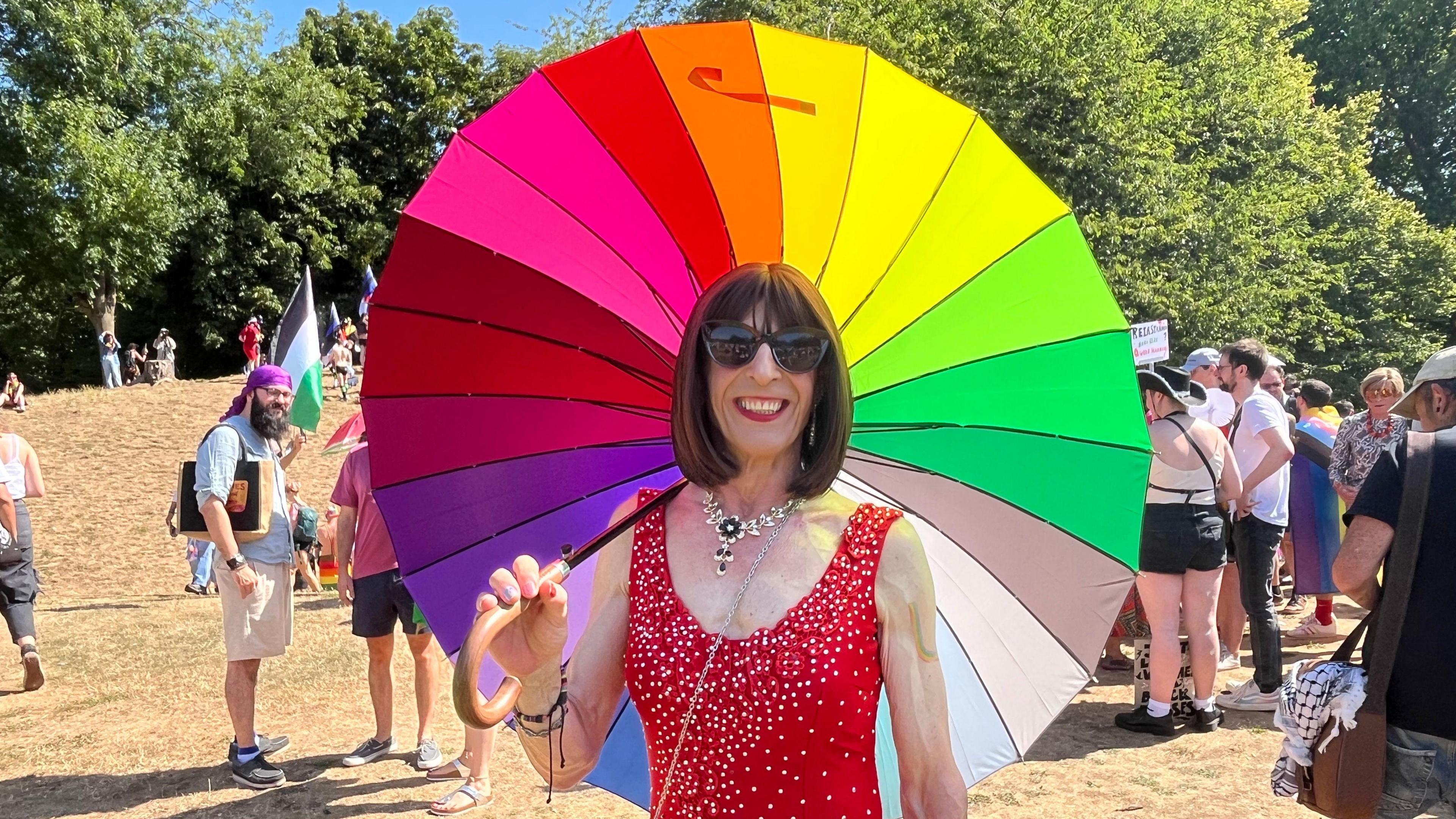 One person holds a huge rainbow umbrella and smiles into the camera. They are wearing large sunglasses and a sparkly red dress. Behind are lots of people in the park, some with flags, and trees behind them