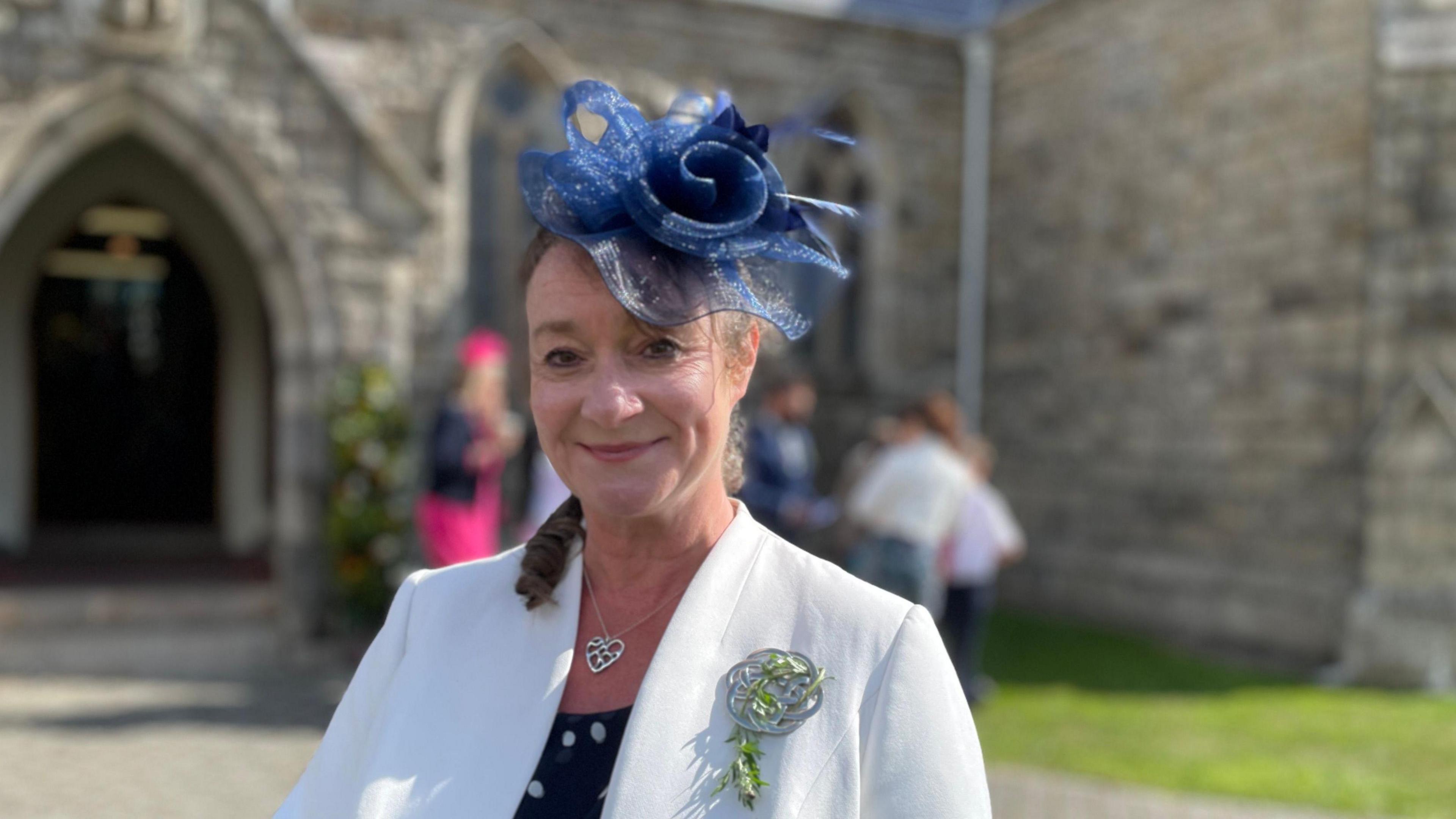 Daphne Caine, a woman with a white blazer and a blue flower hat smiles as she stands in front of St John's Church.