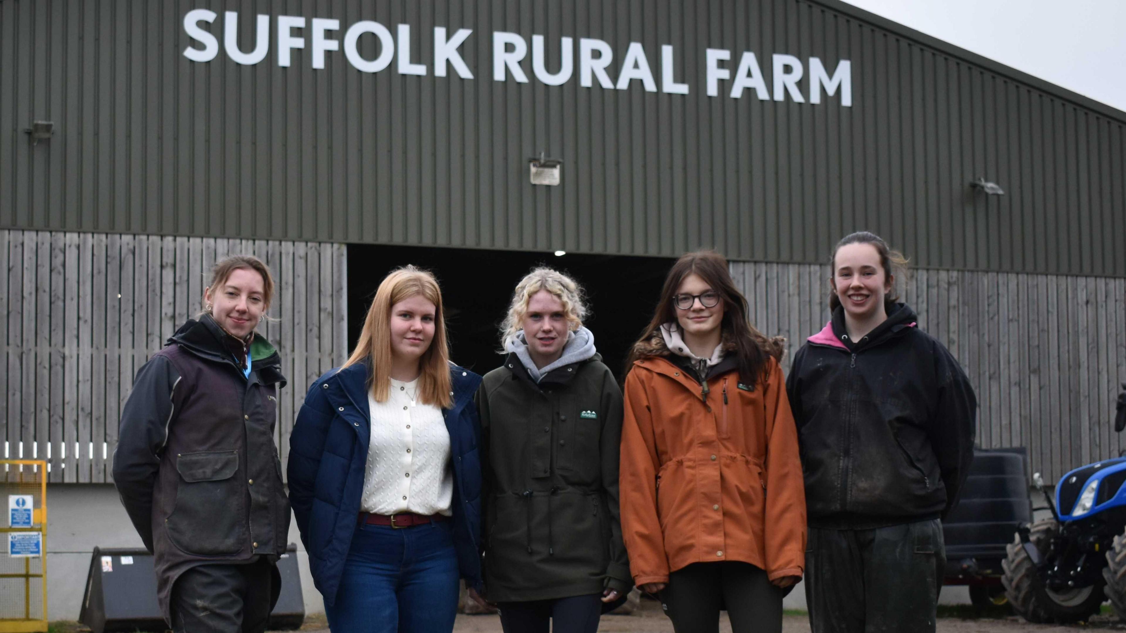 Five women stand in front of a barn which says Suffolk Rural Farm in white capital letters. Enya wears a black waterproof jacket, and has her hair tied back. Daisy has a blue coat and jeans and a white jumper. She has long blonde hair. Ellie has a black coat and blonde hair. Mollie-Mae wears and orange coat with a hood and has long brown hair and glasses. Sophia is wearing a black jacket and jeans and black waterproof trousers. Her brown her is tied back off her face.