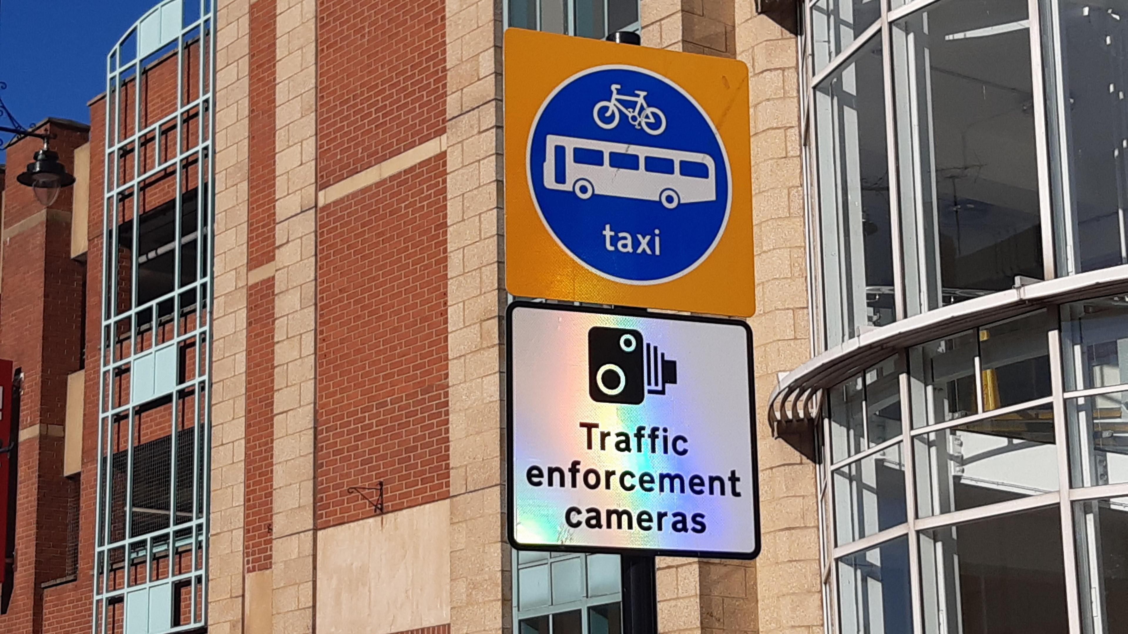 A white square bus gate enforcement sign, reading Traffic enforcement cameras. There is a second sign above it with a picture of a bus, bike and the world taxi written on it.