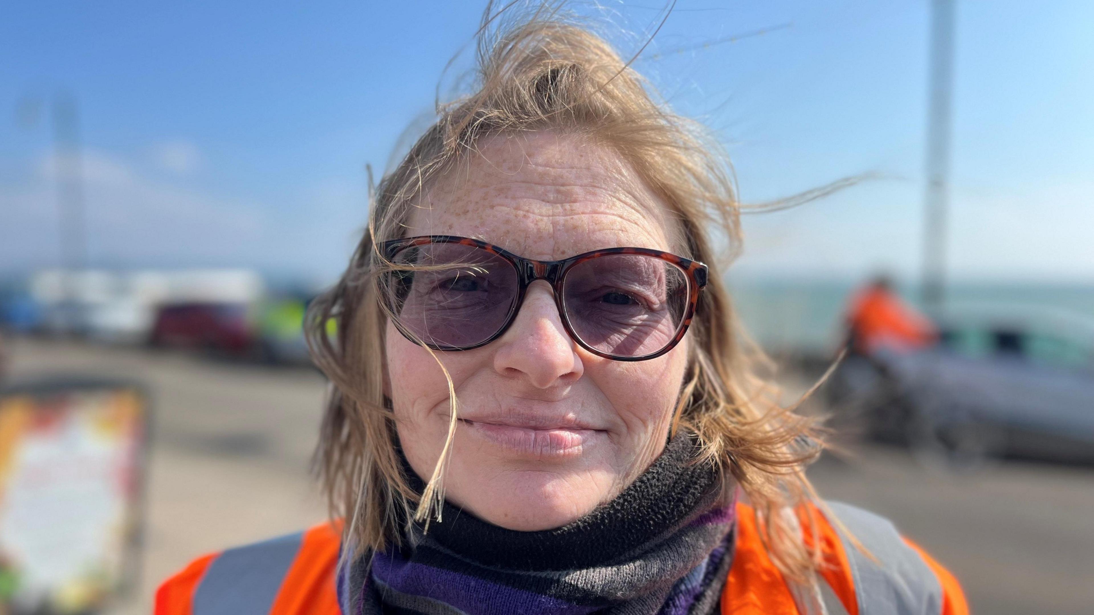 A lady with fair hair that is blowing in the wind, looking straight at the camera, wearing sunglasses and an orange hi-vis jacket, you can see the sea and blue sky in the background.