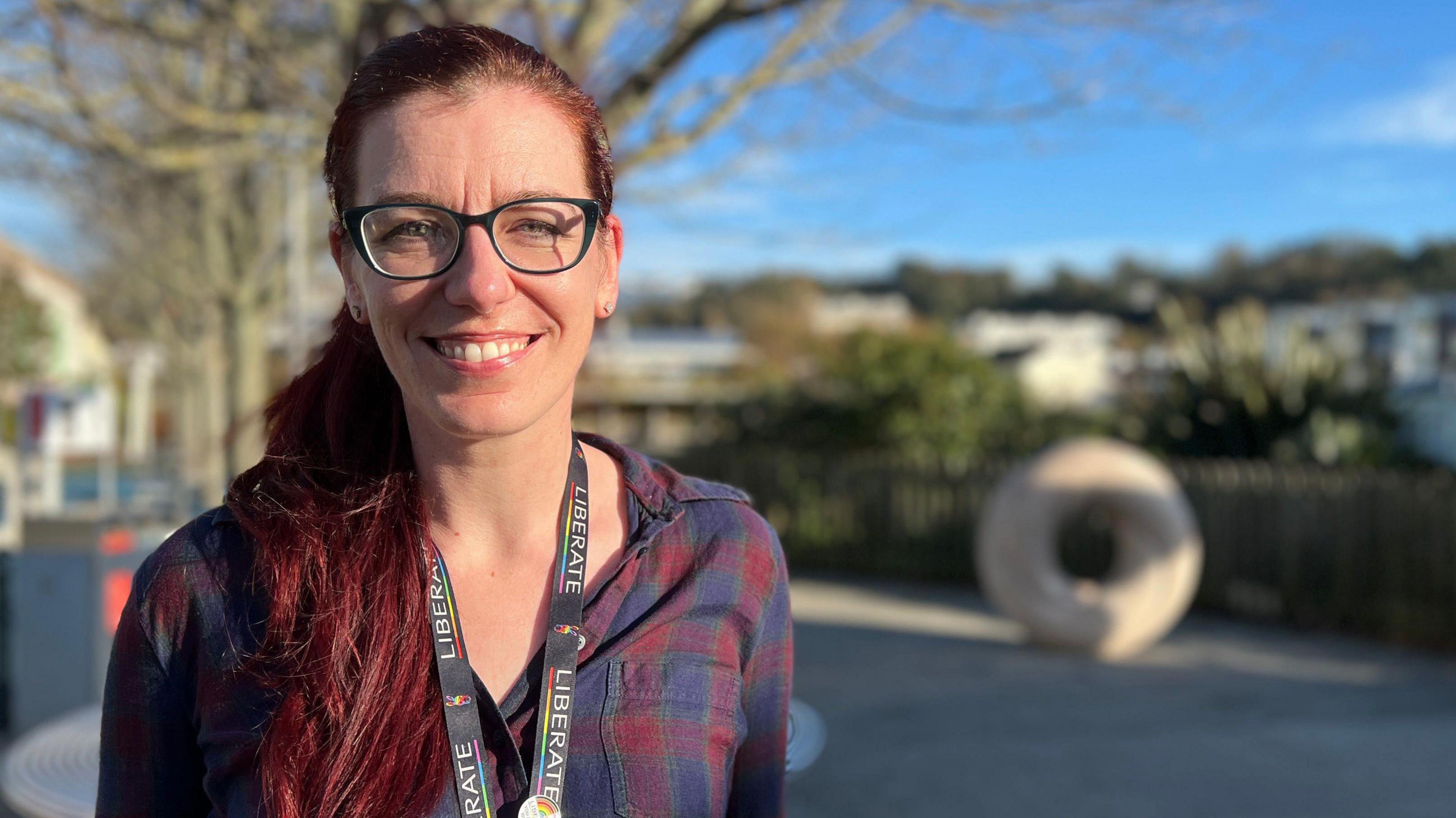 Kaye is standing outdoors in a park-like area with a tree in the background. She is wearing a dark plaid shirt and a lanyard with visible text. Behind her, there is a large circular stone sculpture and a wooden fence, with buildings and greenery in the distance under a clear blue sky.