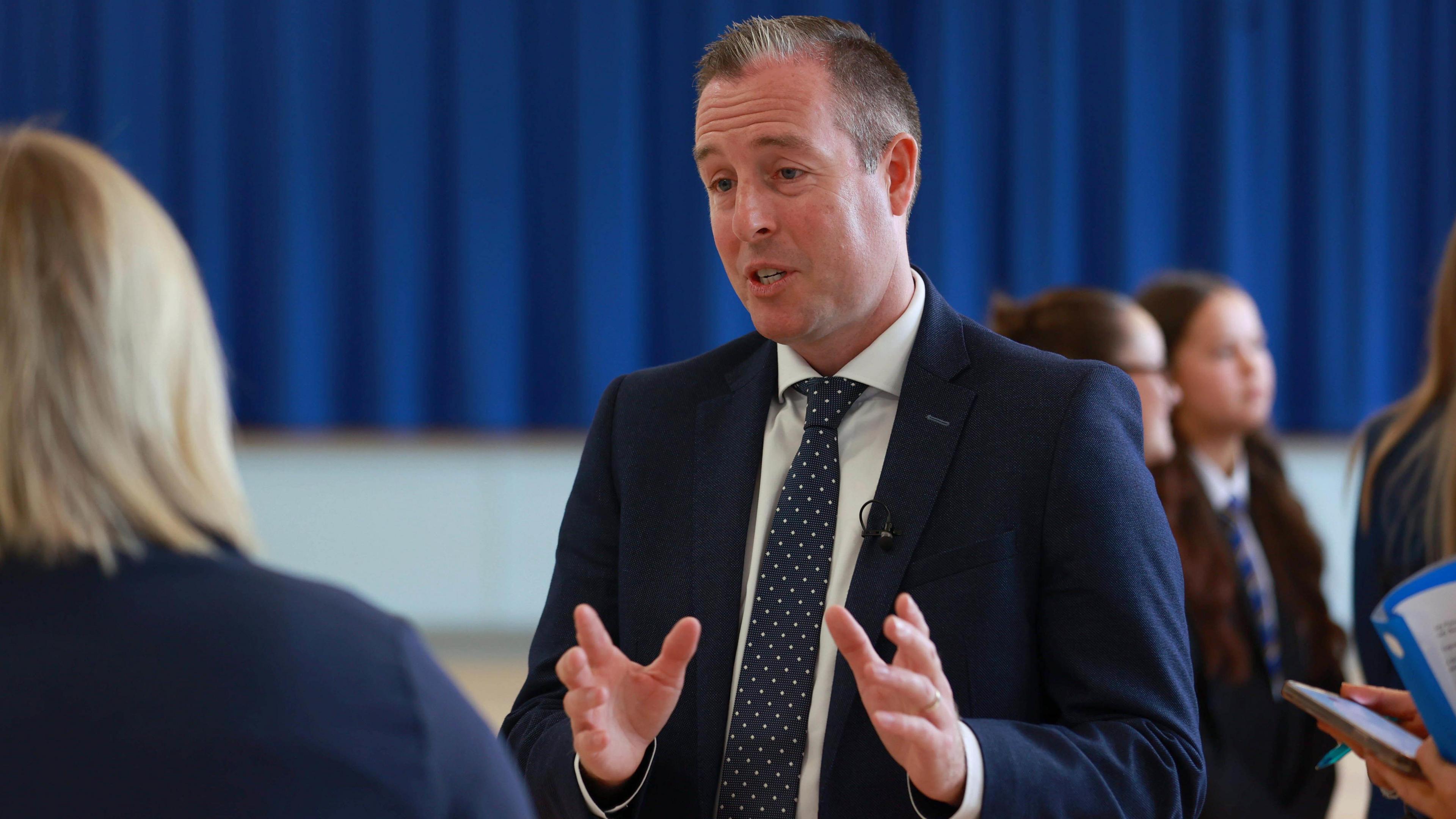 Paul Givan speaking with his hands out in front of him. He is wearing a suit and tie. He is speaking to a woman with her back to the camera. Behind him there are some girls in school uniform and behind them a large blue curtain.