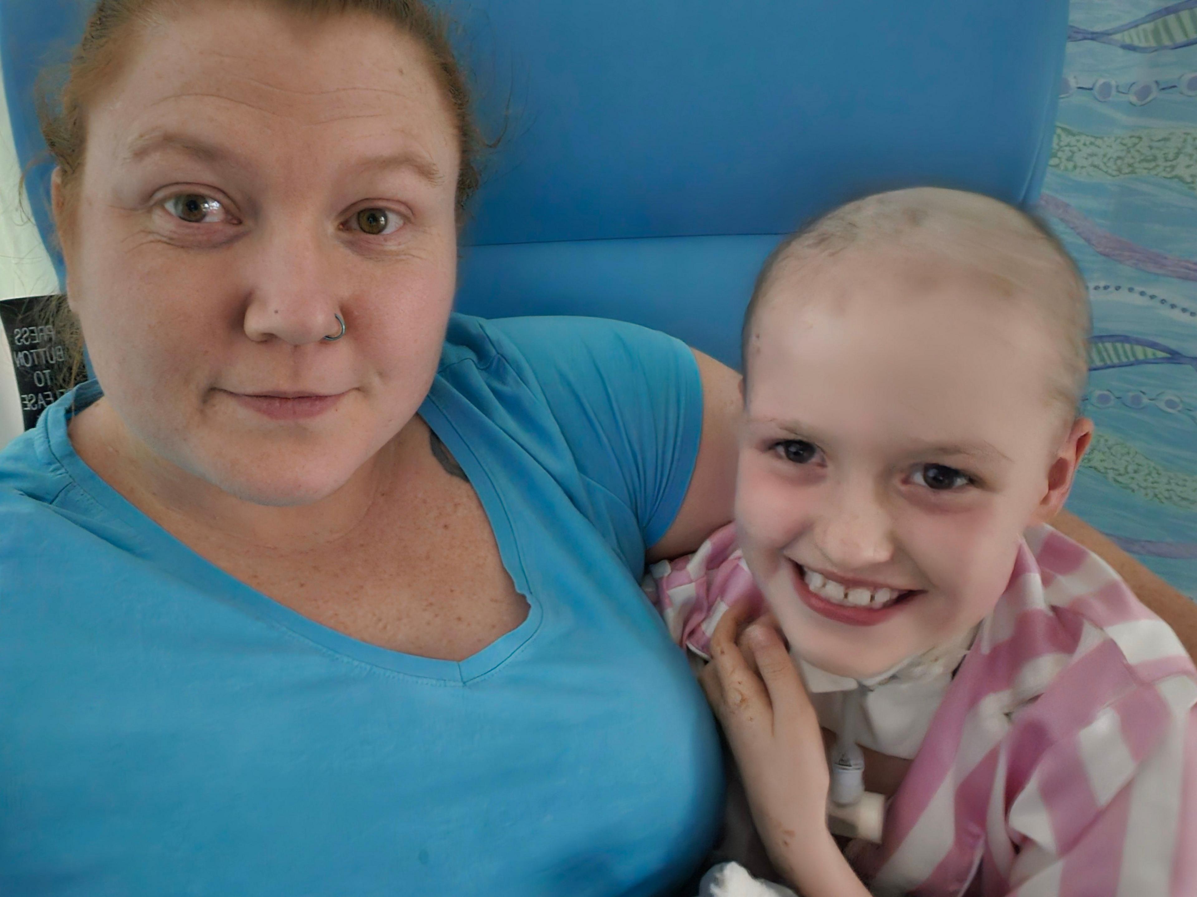 A woman and a young girl hug in a blue chair. The woman has brown hair, and is wearing a nose ring and a blue T-shirt. The girl has no hair and is wearing a pink and white striped top. She has a small plastic tube and device attached to the front of her neck.