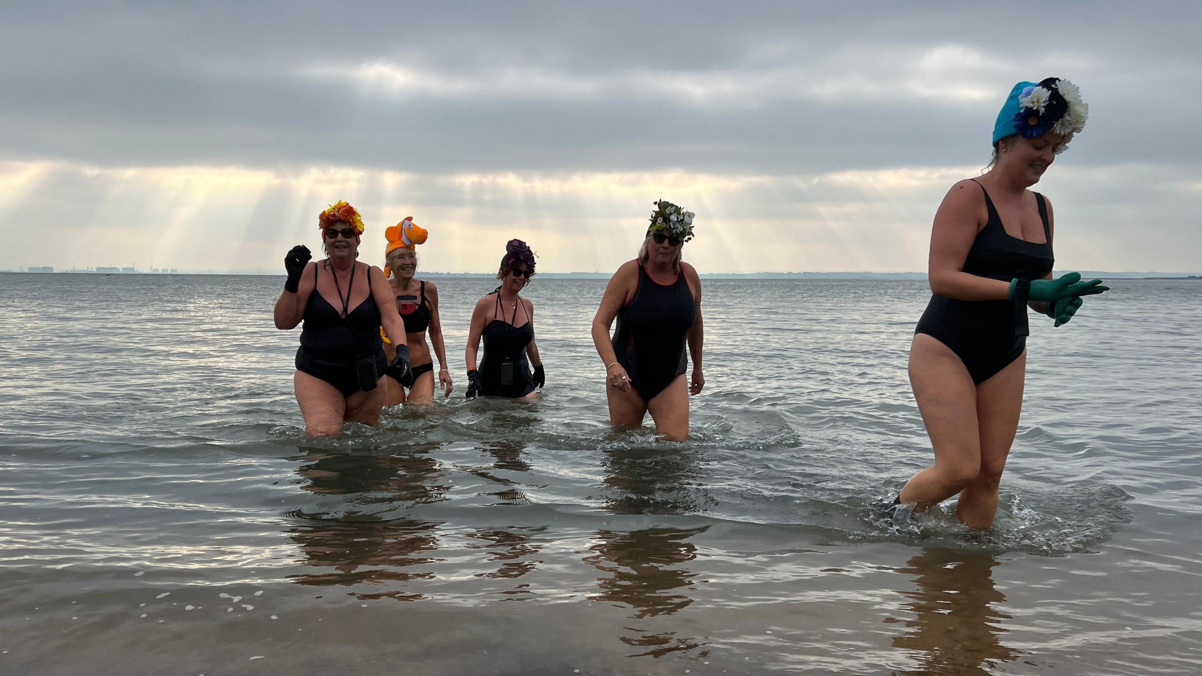 Five women walk out of the sea, four are wearing swimsuits and one is in a bikini. Three of them are wearing flower head-dresses, and another three are wearing gloves. You can see the sun's rays poking through the clouds behind them