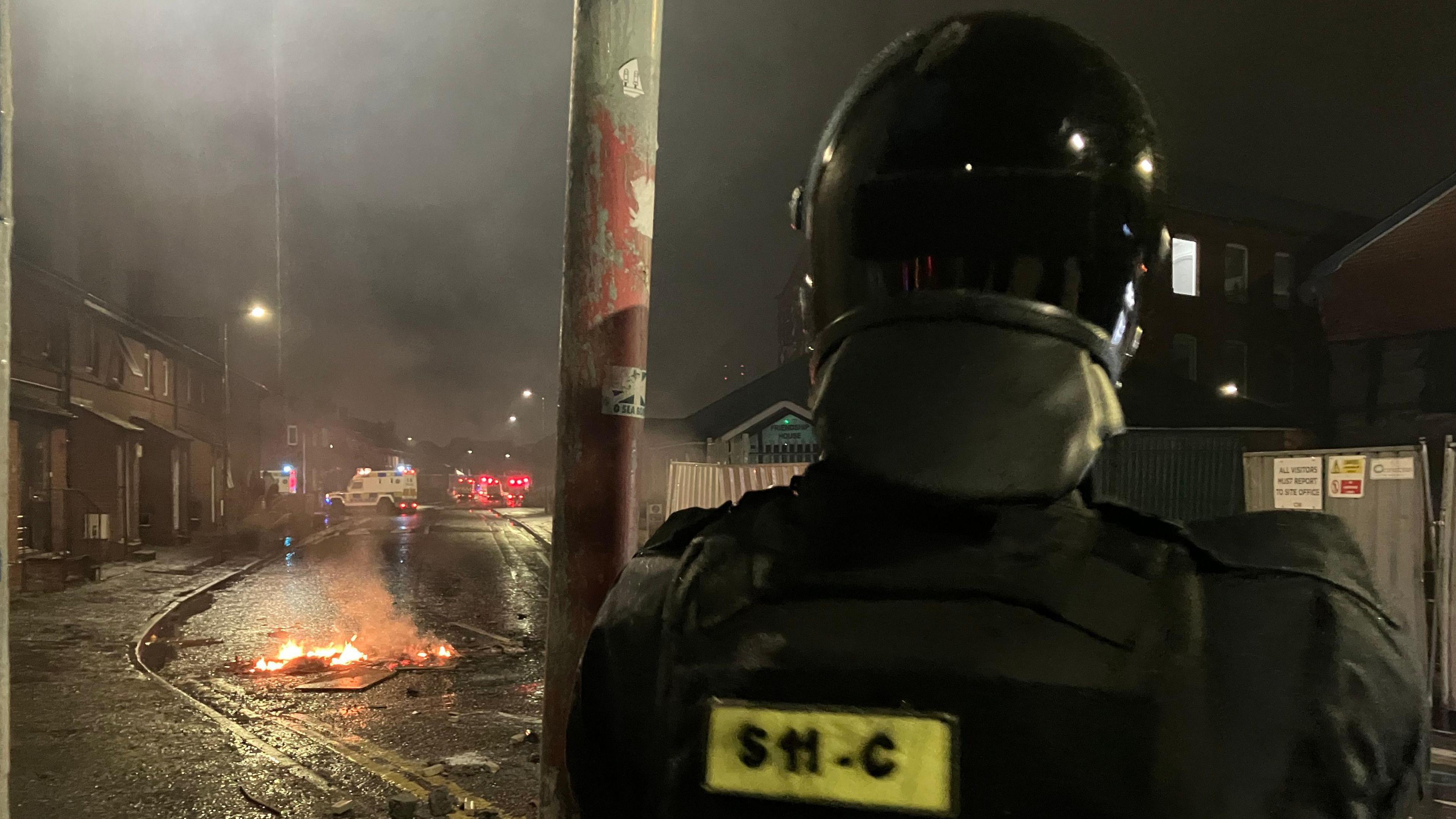 Police officer in riot gear standing looking at something burning in the middle of the road