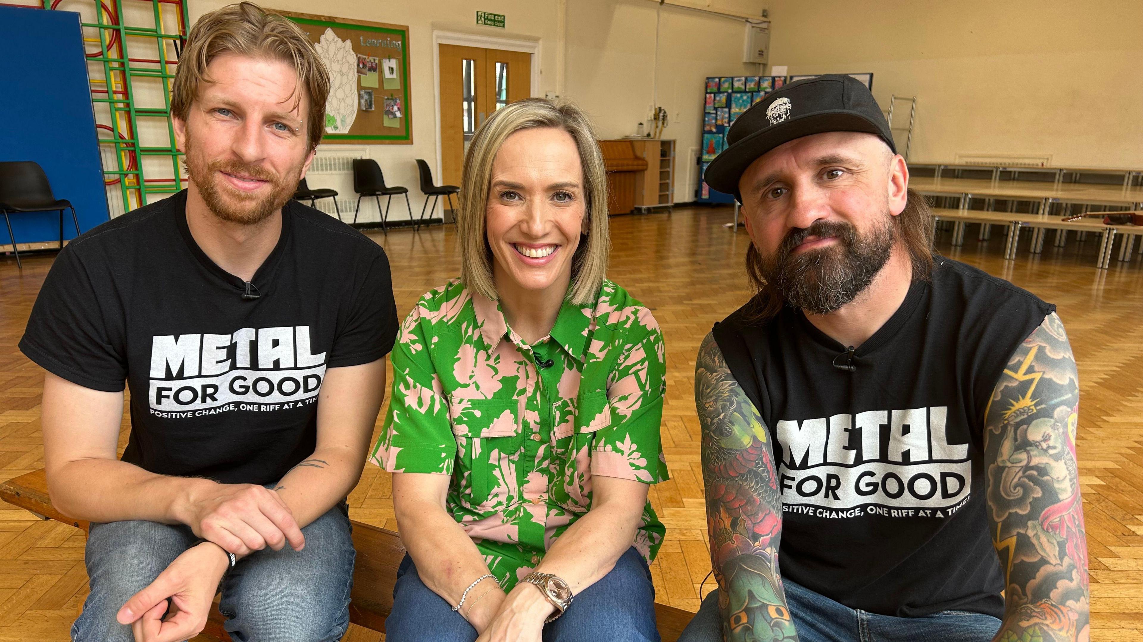 Adam Thistlethwaite and Baz Mills are sitting in the middle of a hall at Trumacar Community Primary School on a bench. They are both wearing black T-shirts which feature the message "Metal for Good". Kay Crewdson is sitting in between them and they are all smiling at the camera 