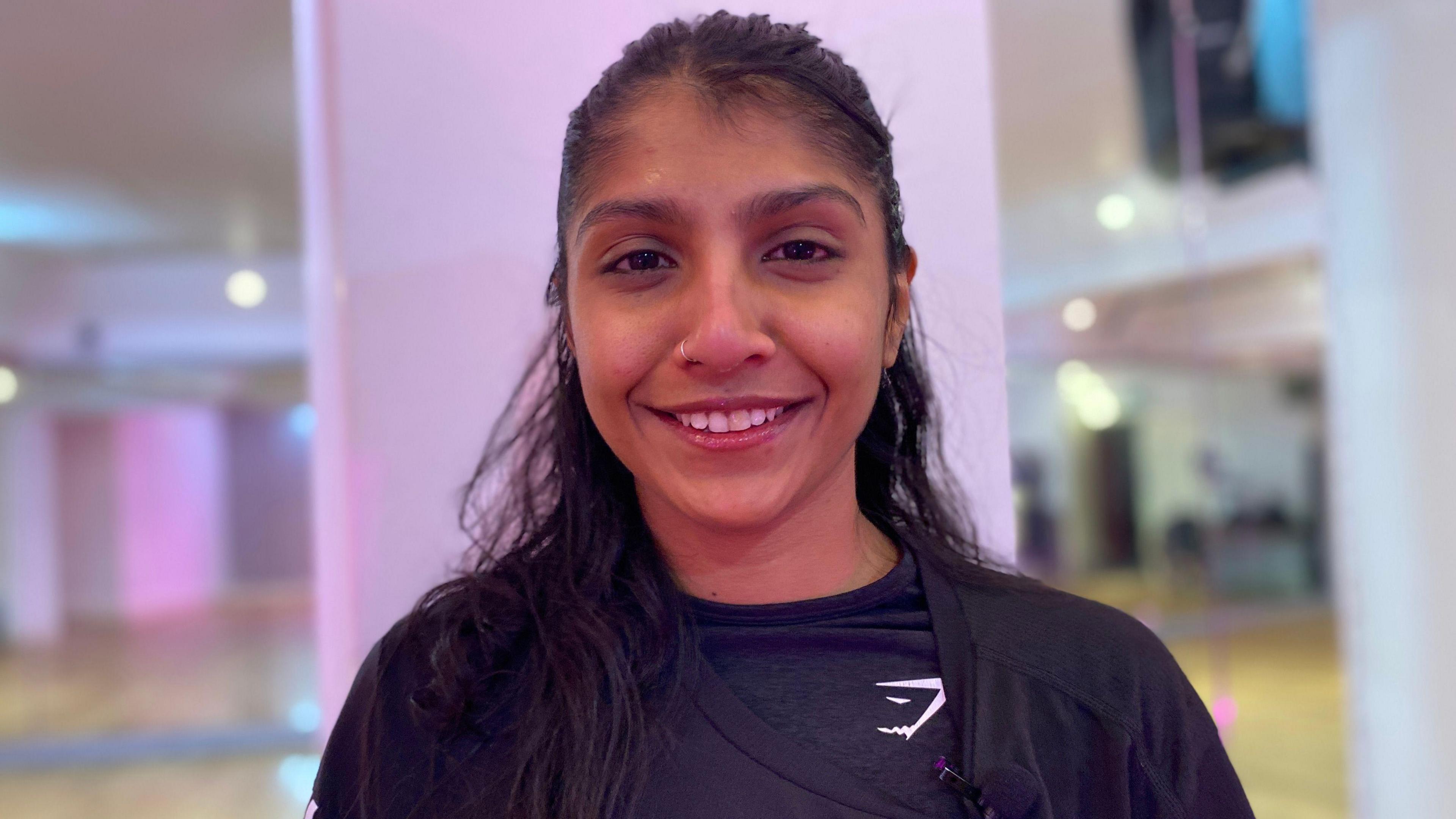 Nishi Panicker is wearing a long-sleeved black gym top and has long brown hair. She is smiling in a gym studio.