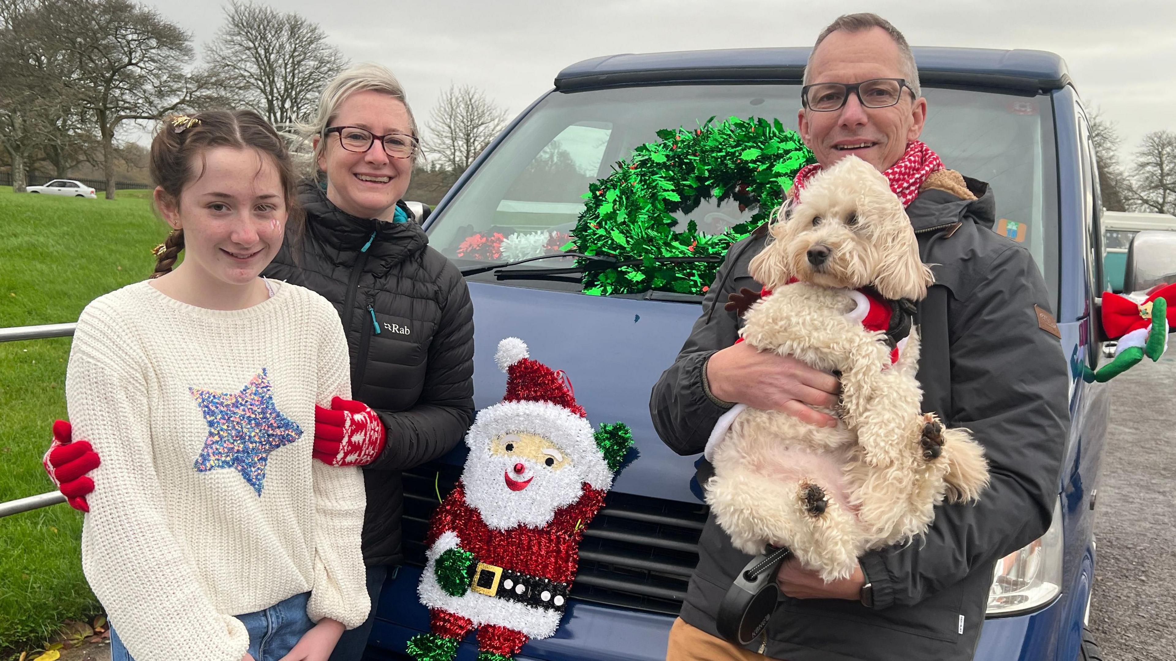 To the right of the picture is the event organiser Phill Ashby. He is leaning against his blue VW van holding his white dog. Standing to on the left of the picture is Phill's wife and daughter. The daughter is dressed in a white jumper with a blue star on it. The van is decorated with a green holly wreath and there is a sparkly Santa Claus on the bonnet.