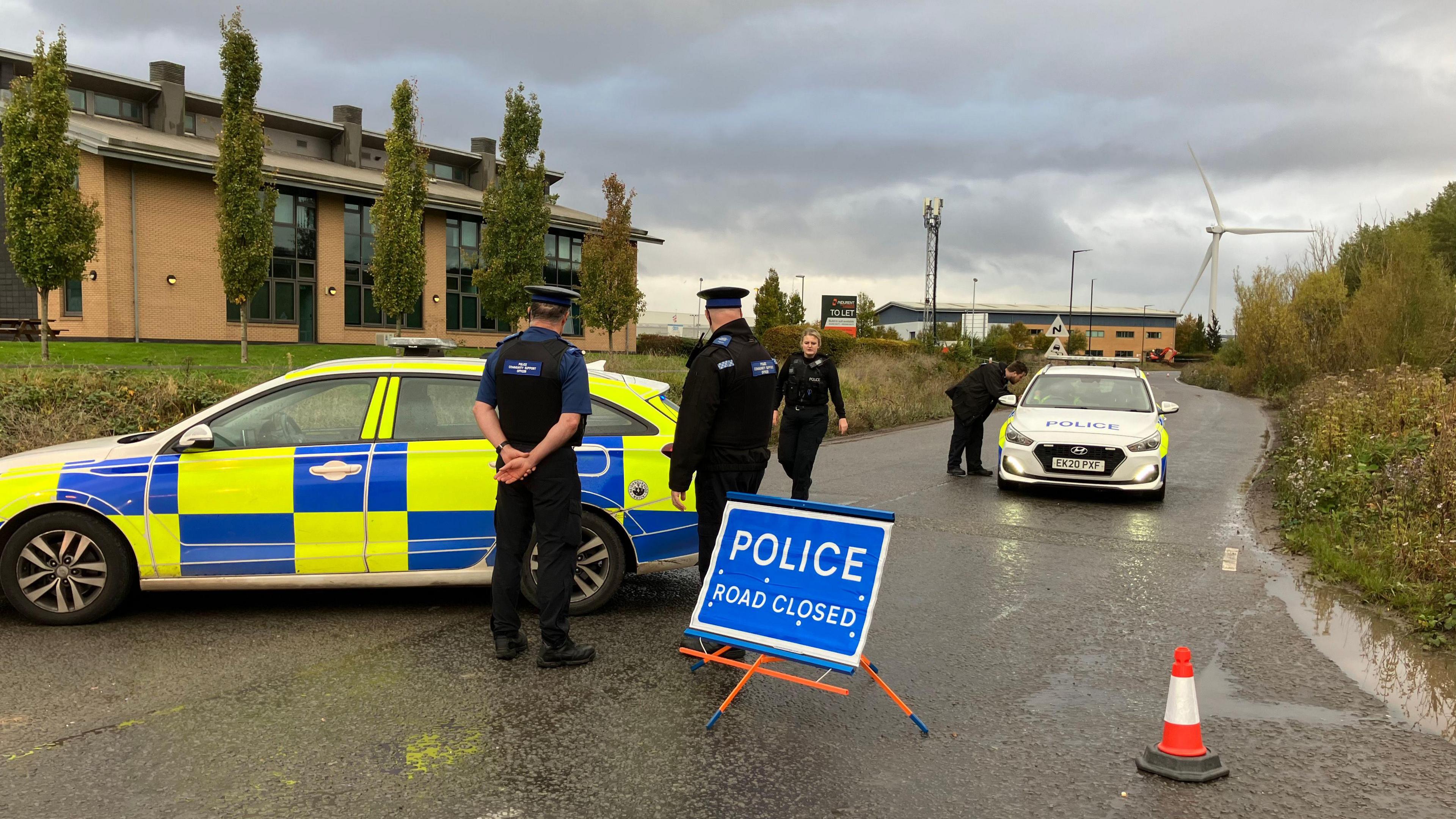 Two police cars and four police officers are standing on a road with a 'police road closed' sign.