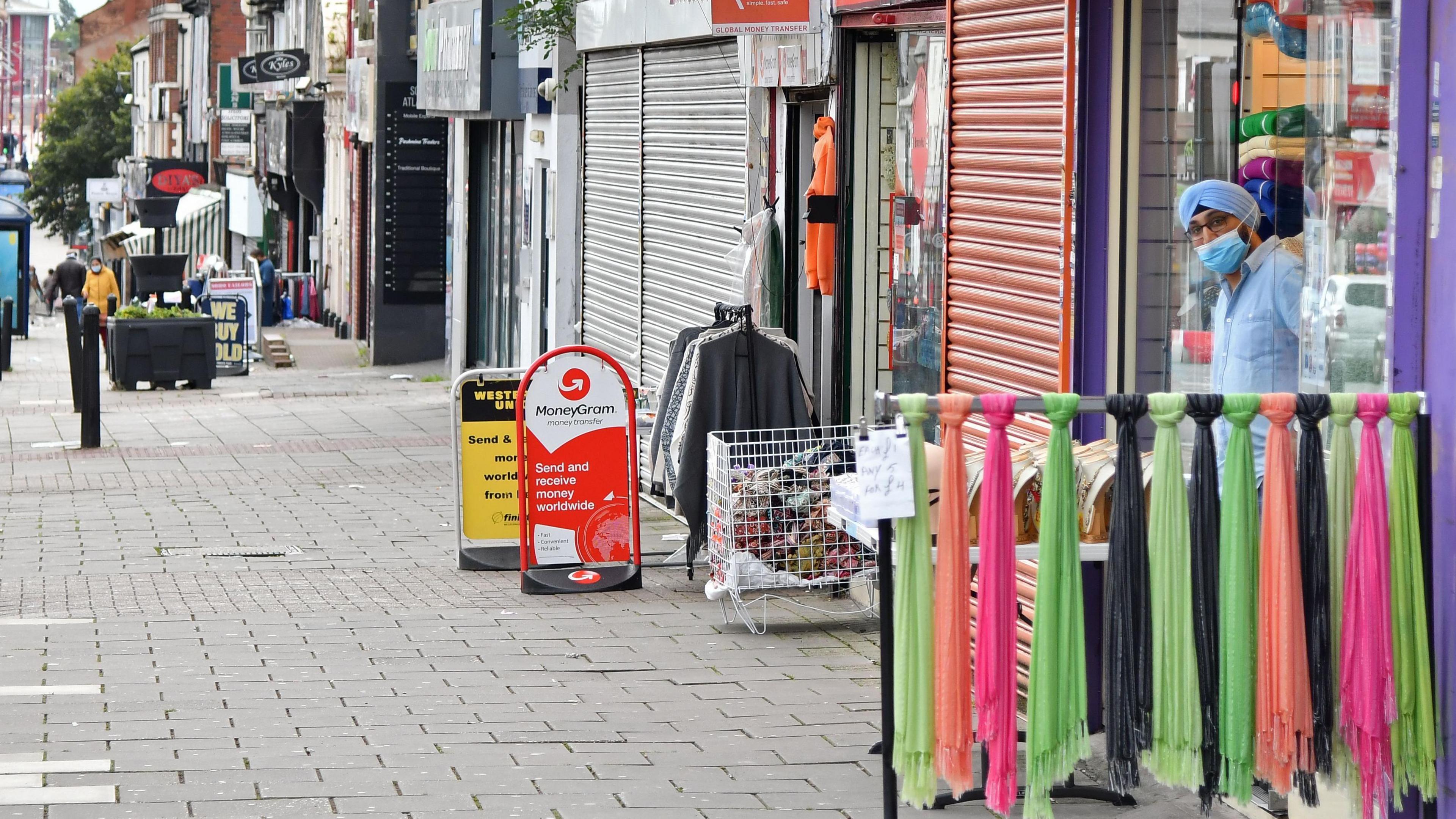 A shot of Soho Road's shops, lined on the right hand side. A man in a turban looks out from a shop door, and in front is a row of colourful scarves