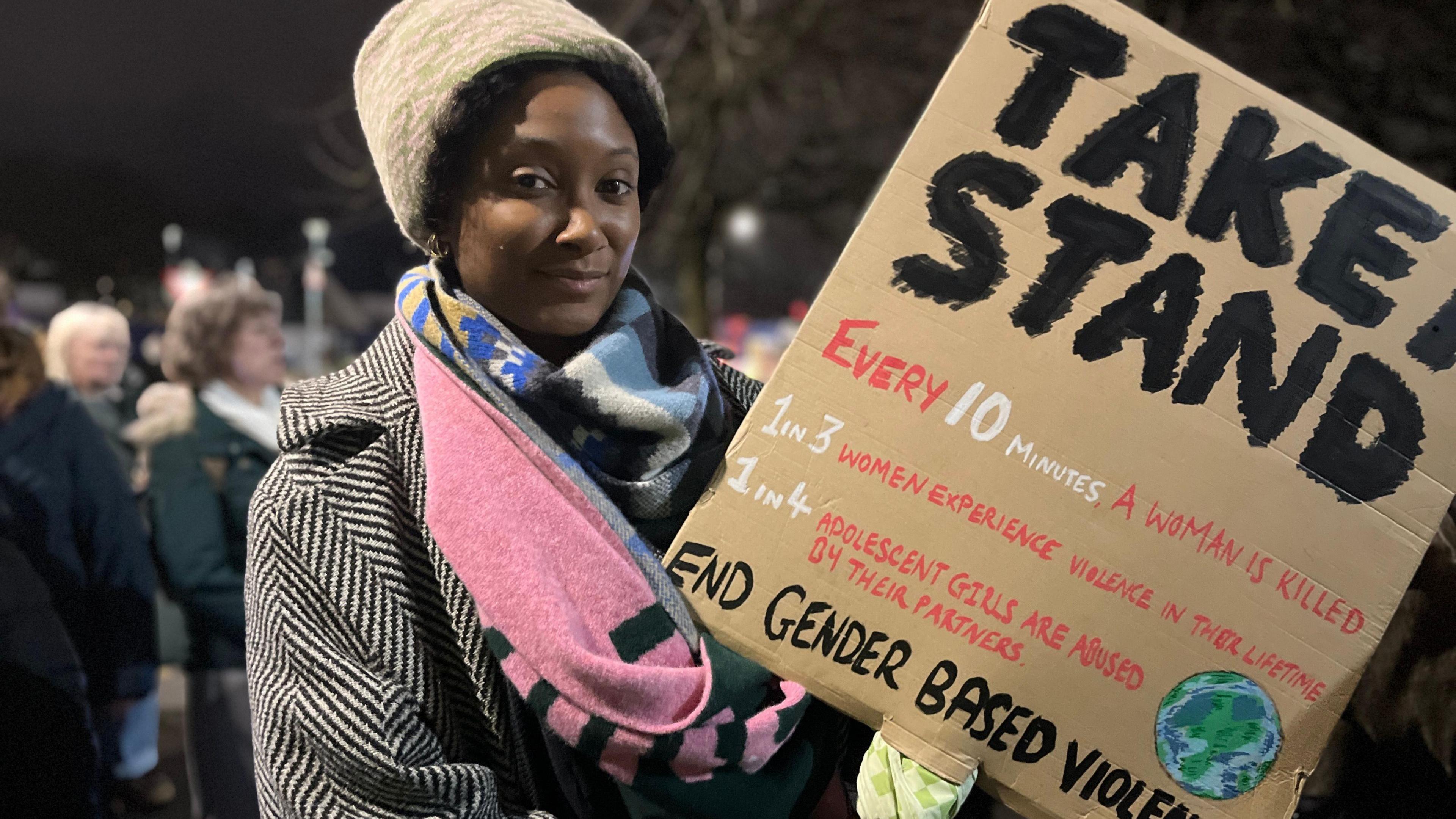 A woman holds a cardboard placard which reads Take a Stand. there is a crowd behind her.