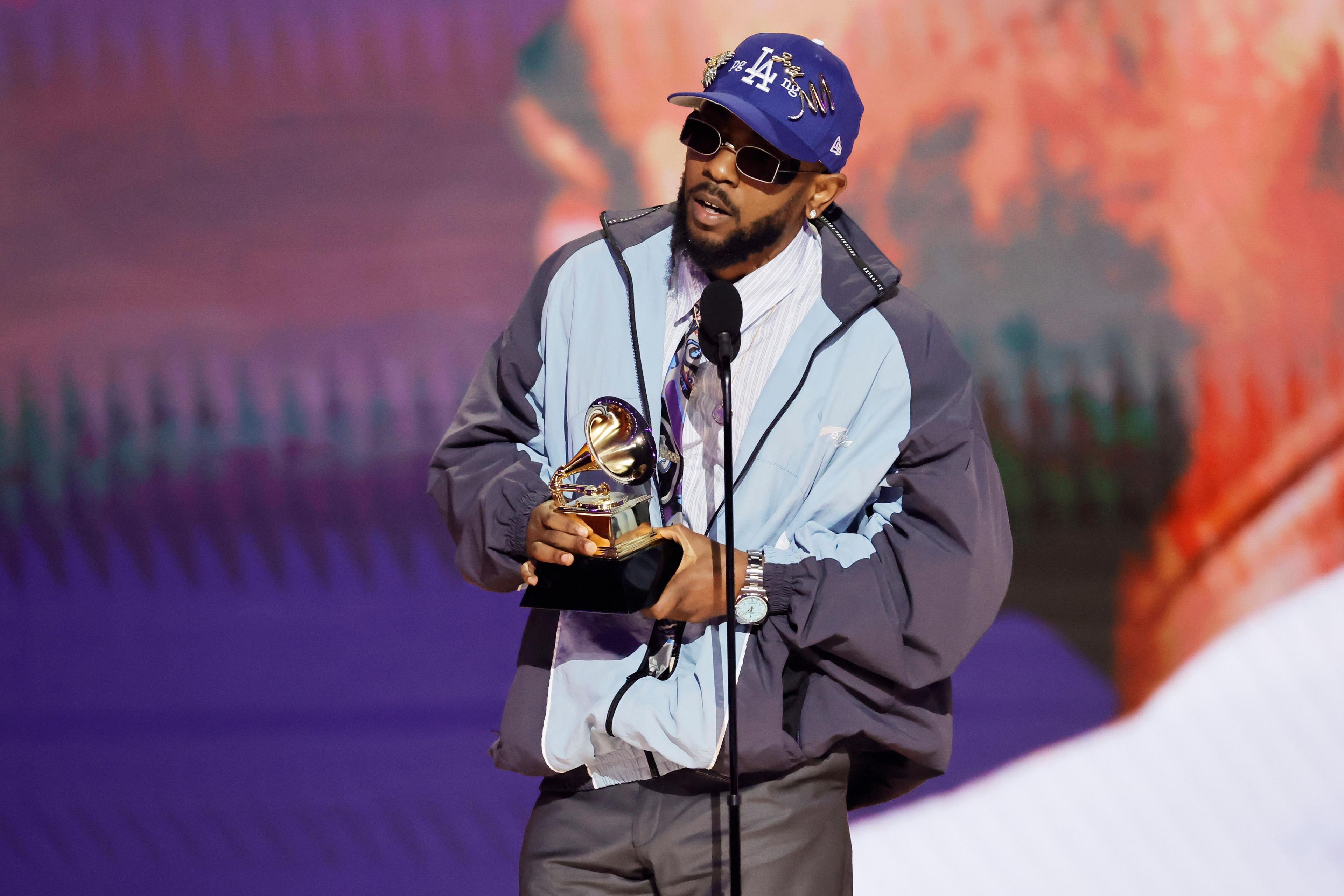 Kendrick Lamar accepts a Grammy Award in 2023. He is wearing a light sports jacket over a suit and tie, with a purple "Los Angeles" baseball cap and blackout sunglasses.