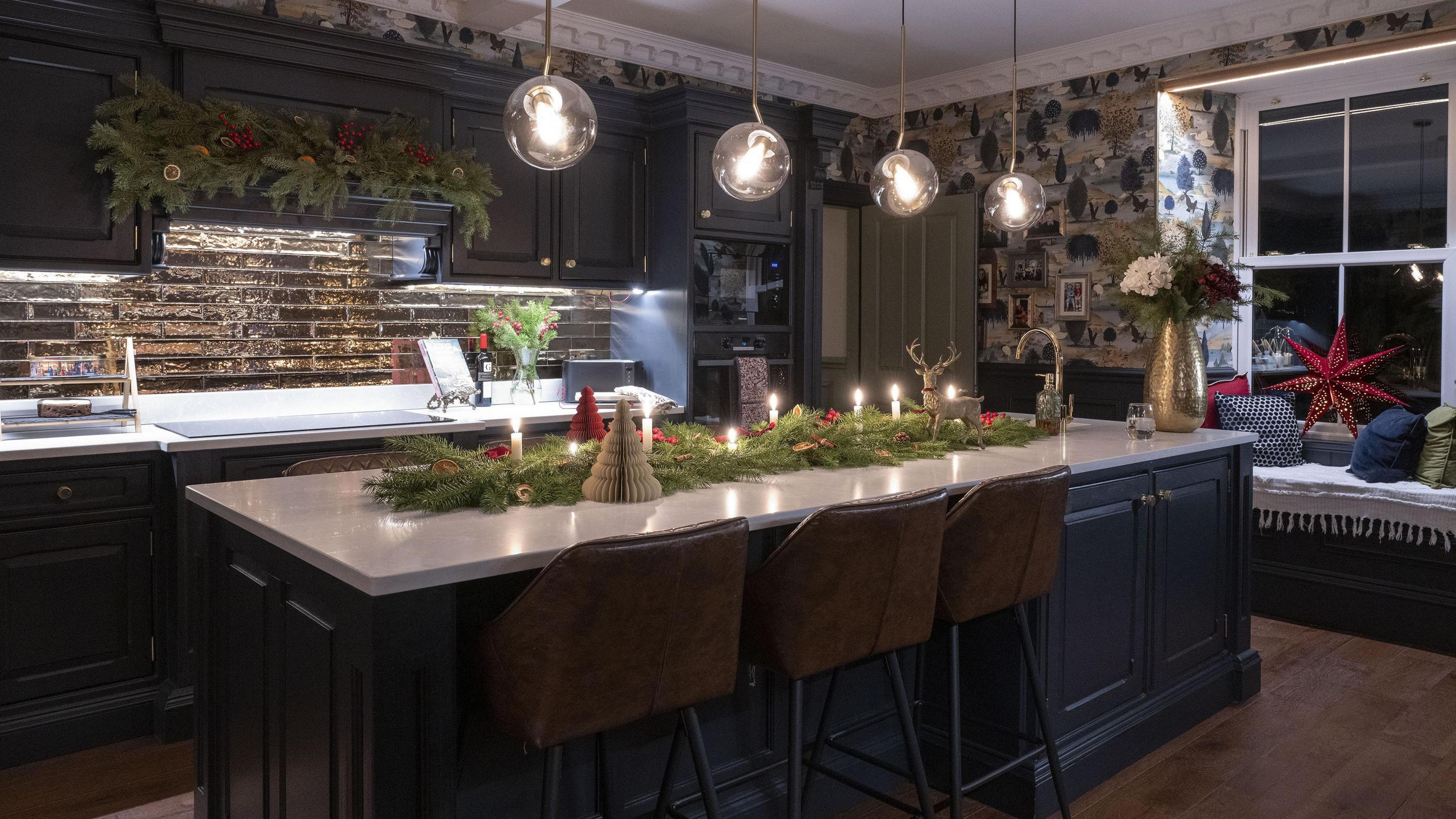 A kitchen with cupboards made of navy wooden panelling. Christmas themed foliage, with lit candles in it, lies atop a large breakfast bar in the middle of the room. A garland  is also hung above the hob. 