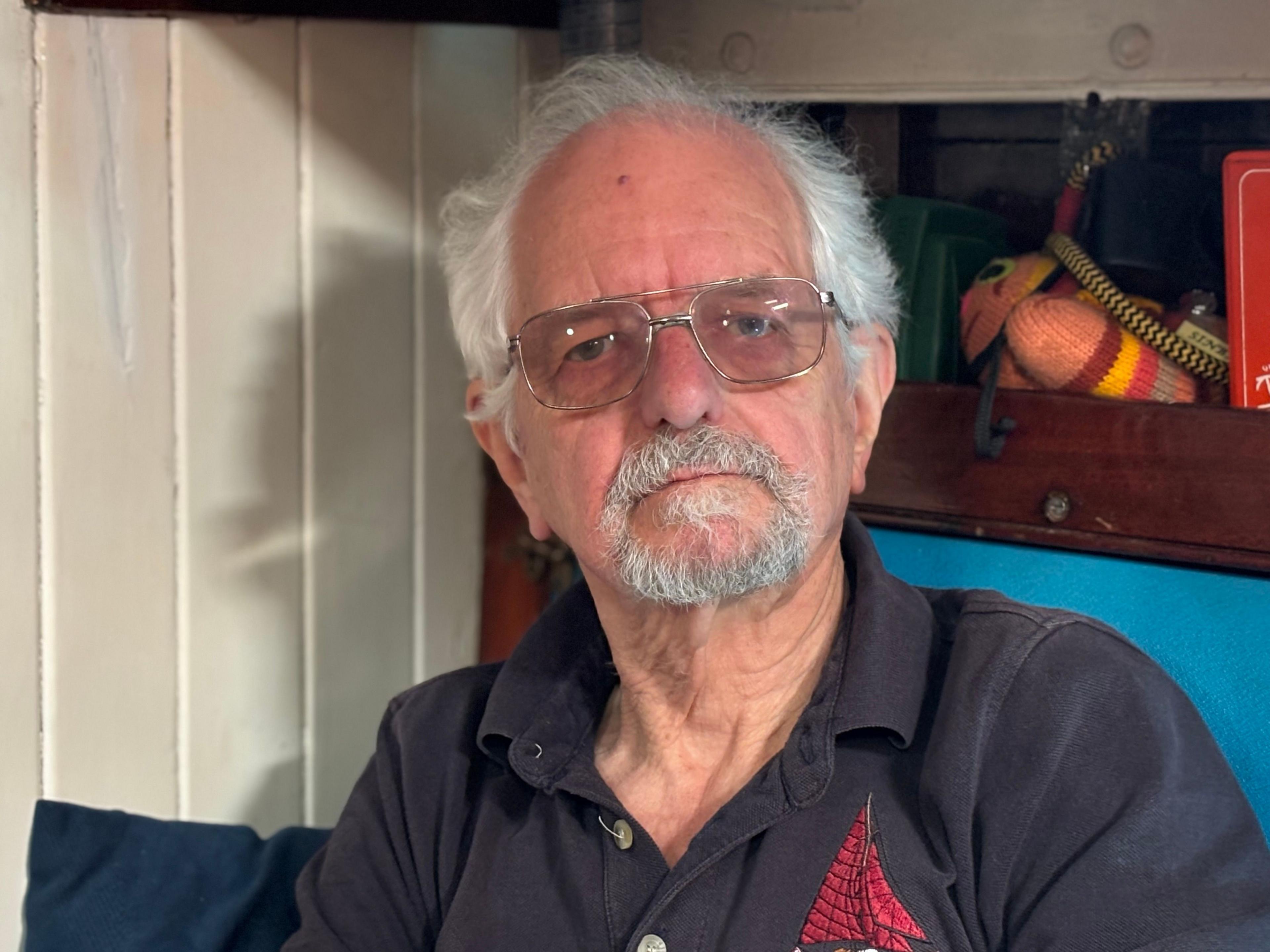 An older man with white hair, a short beard, and glasses sits indoors against a wooden panelled wall. He is wearing a dark polo shirt embroidered with a red sailboat design. Behind him are shelves with books and nautical items.