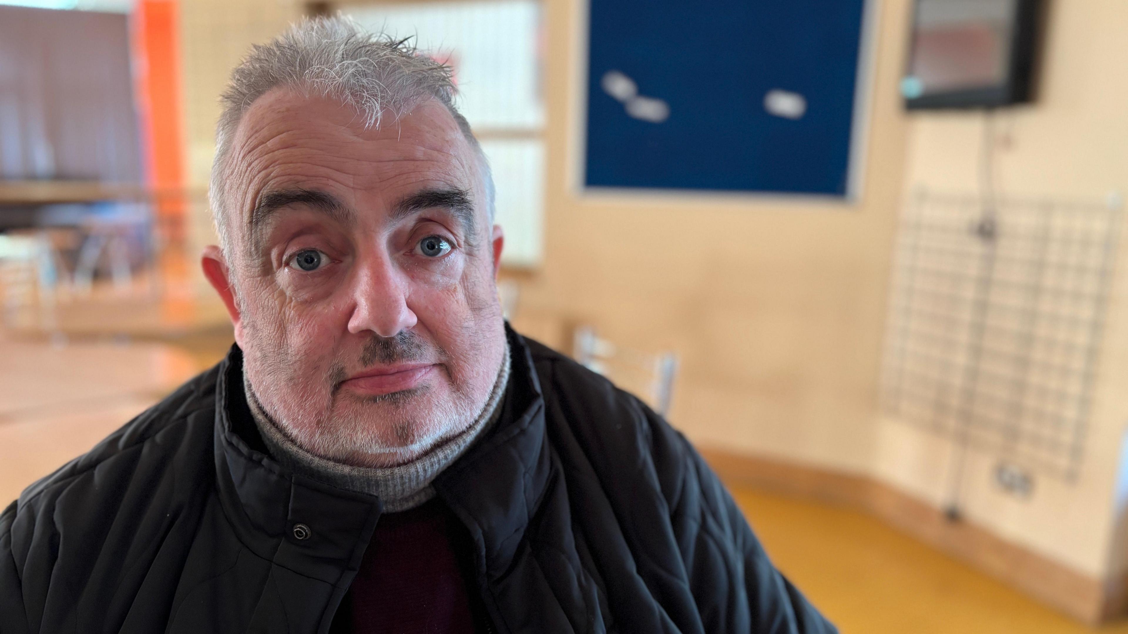 A man sits in a leisure centre. He is dressed warmly in a black coat with a grey woolen polo neck jumper underneath. He has grey hair and dark eyebrows.