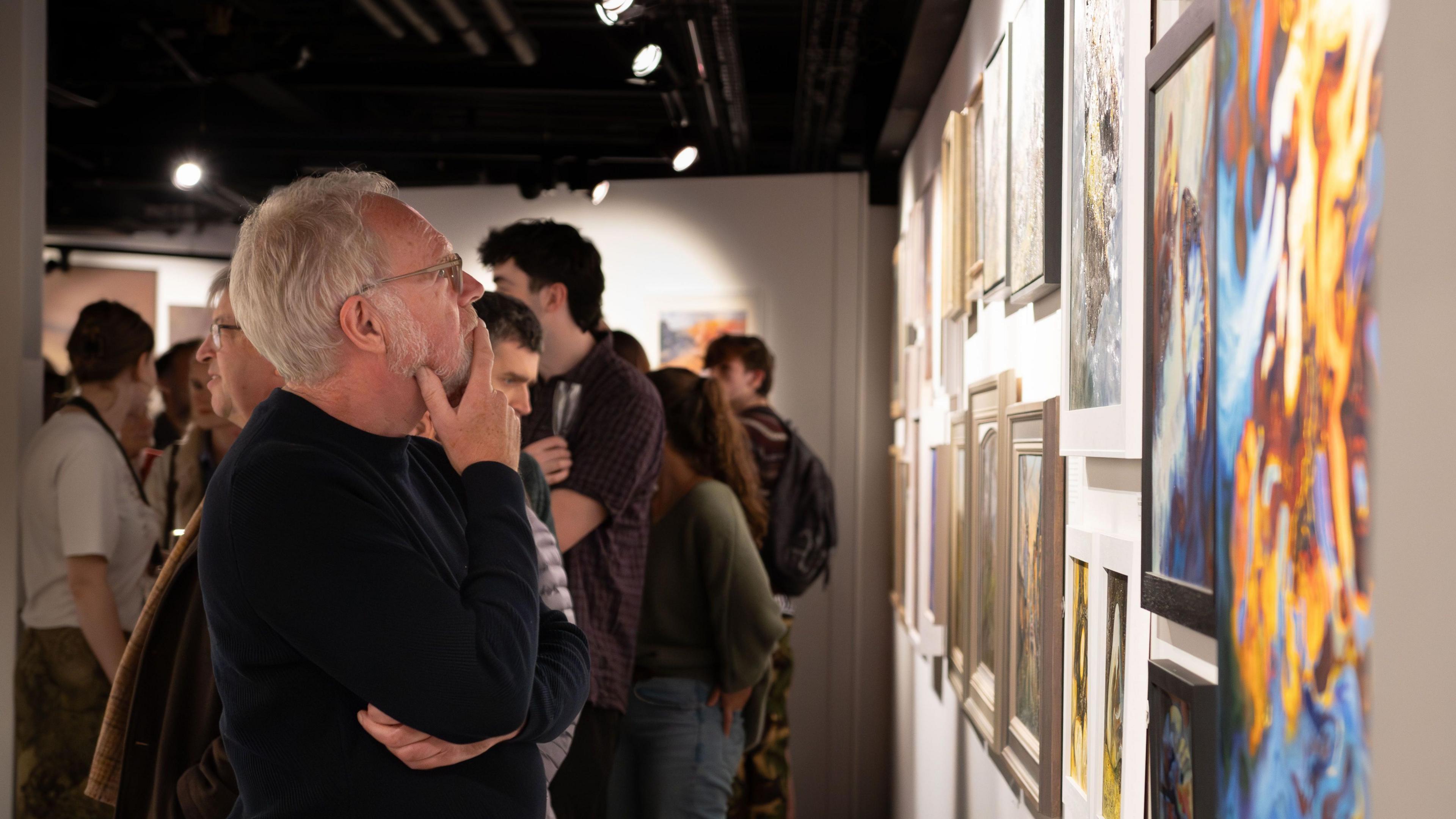 A man with grey hair, glasses and a dark jumper looks at the paintings on one of the gallery's walls. Behind him lots of people are milling around the gallery space.