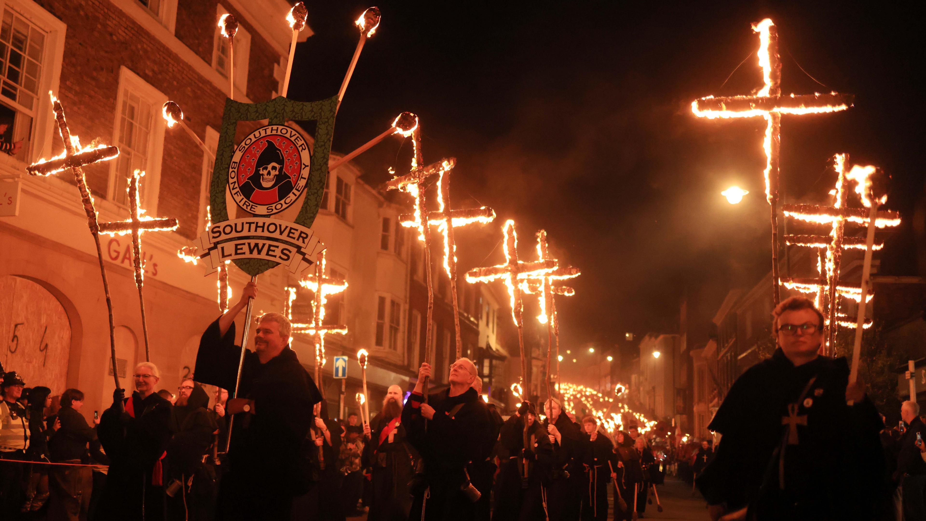 A torchlit procession with flaming crosses in the foreground which is seen stretching back along a long street and with a banner in the foreground which holds the words Southover Bonfire Society and a picture of a skeleton in a black hooded cape.